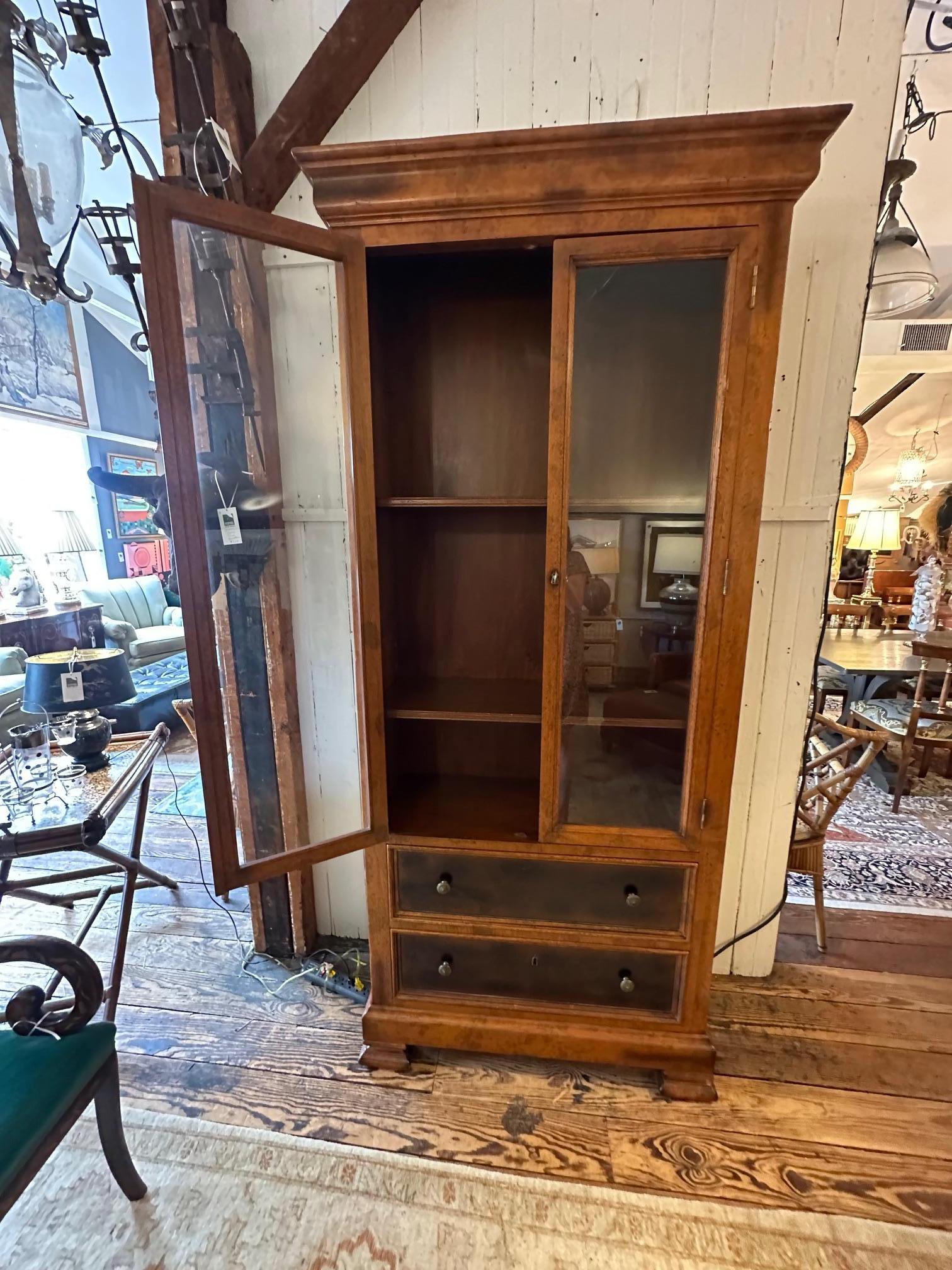 Provincial bibliotheque bookcase cabinet having earthy warm  maple wood with glass front doors and original brass hardware.  Two lower drawers are a darker shade and there are adjustable shelves.