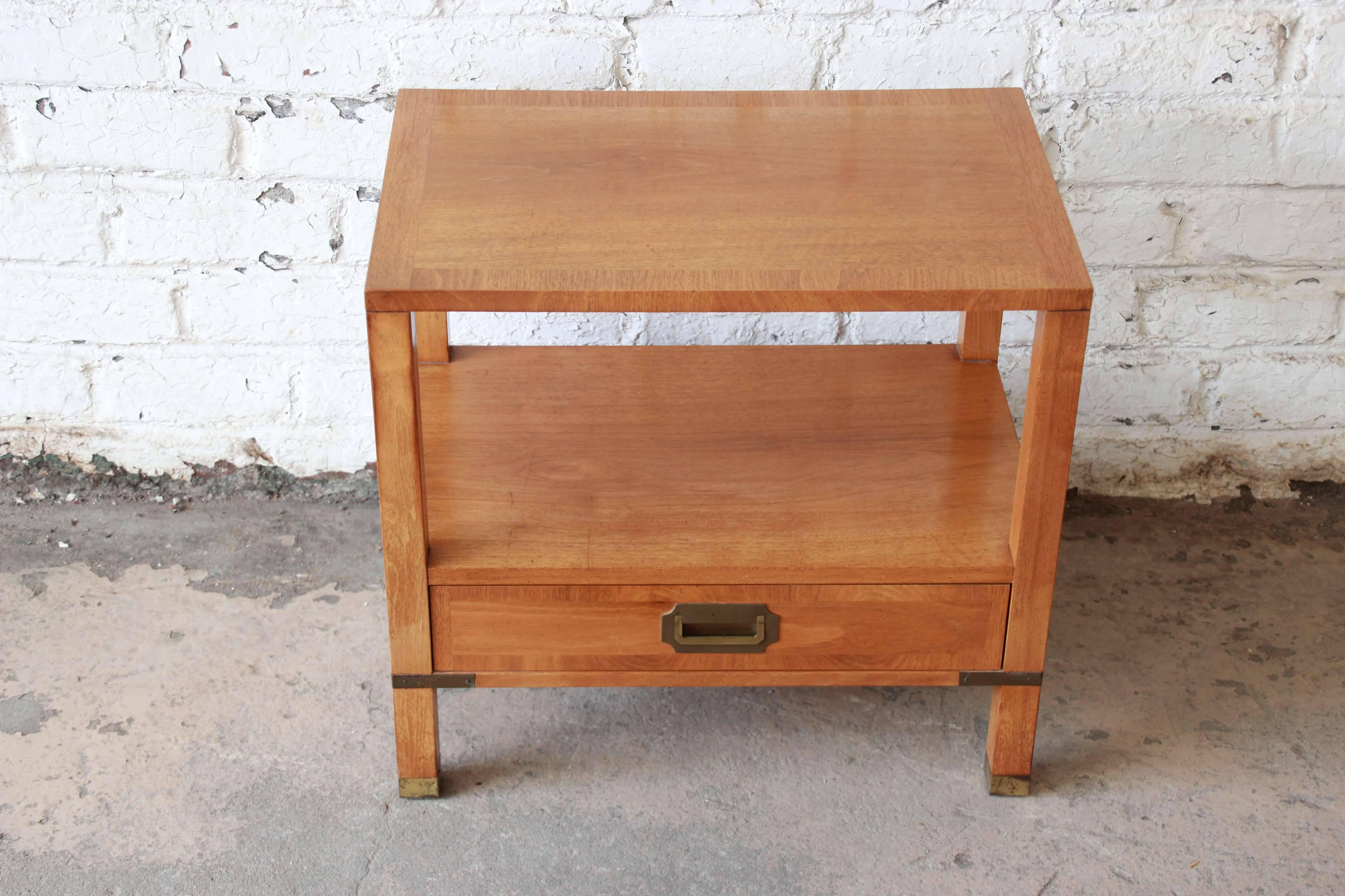 Offering a very nice Baker Furniture Milling Road midcentury campaign style nightstand or end table. The stand has brass hardware and decor in a modern version of this style. There are two tiers for storage with a drawer below. The piece is in good