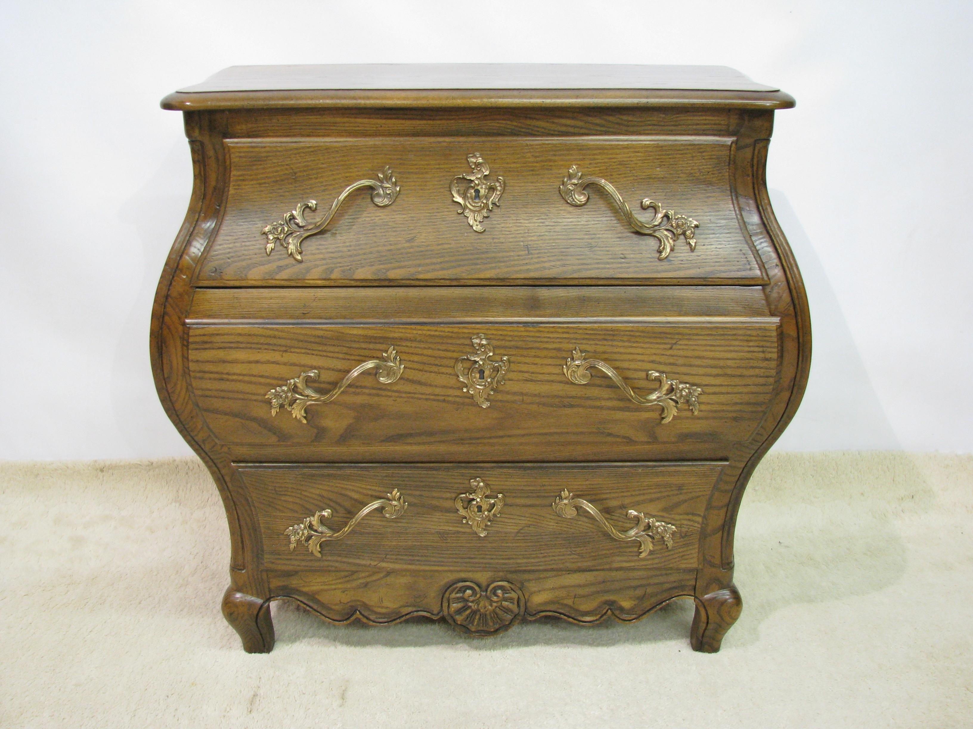 Elegant small scale Bombe chest by Baker Furniture. Solid oak, with a shaped top over a dramatically curved front. Medium brown satin finish, with solid antiqued brass Louis XV style hardware. Solid oak secondary woods and dovetailed drawers. Baker