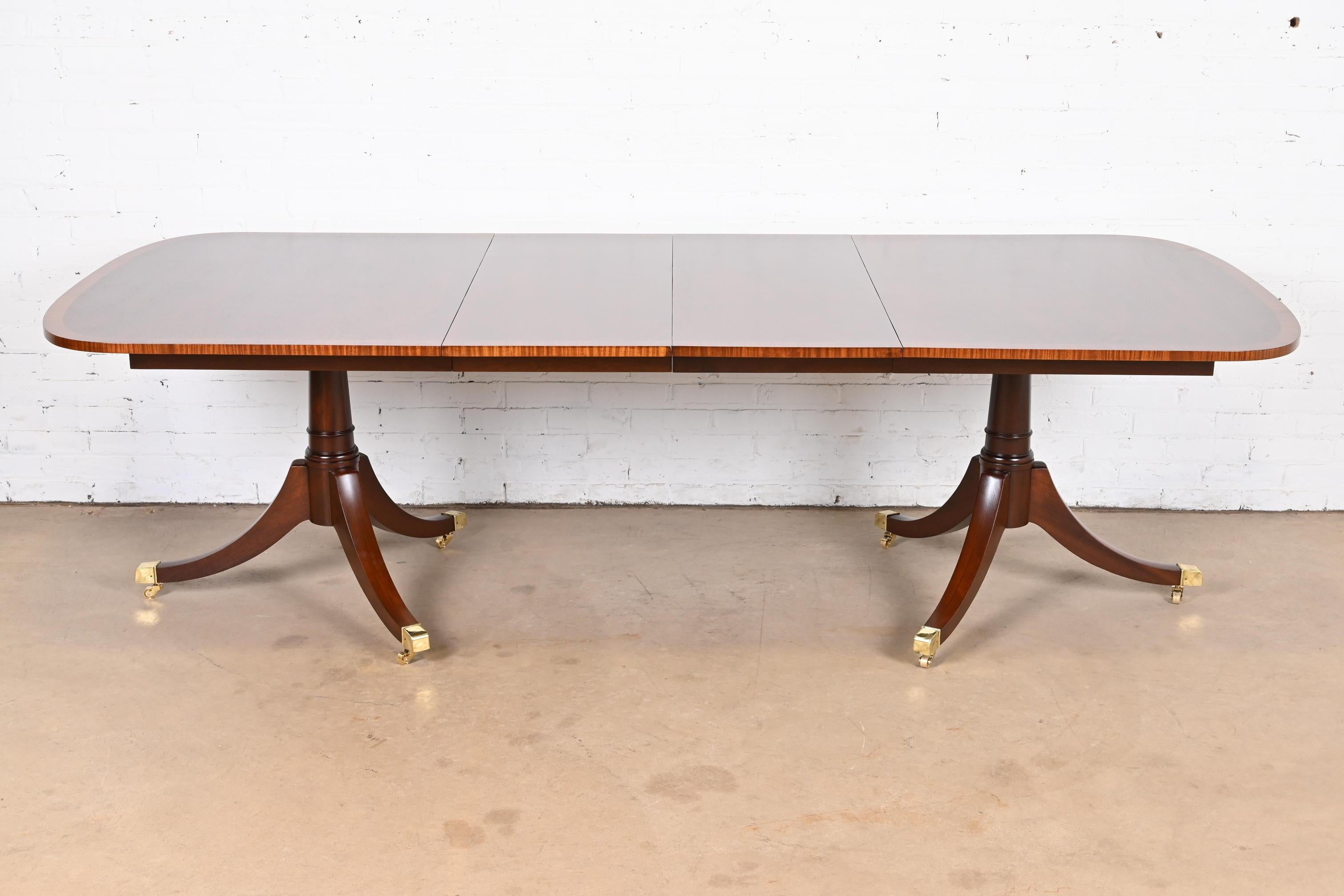 An exceptional Georgian or Regency style double pedestal extension dining table

In the manner of Baker Furniture

USA, Circa 1980s

Gorgeous mahogany top, with satinwood banding, carved mahogany pedestals, and brass capped feet on brass