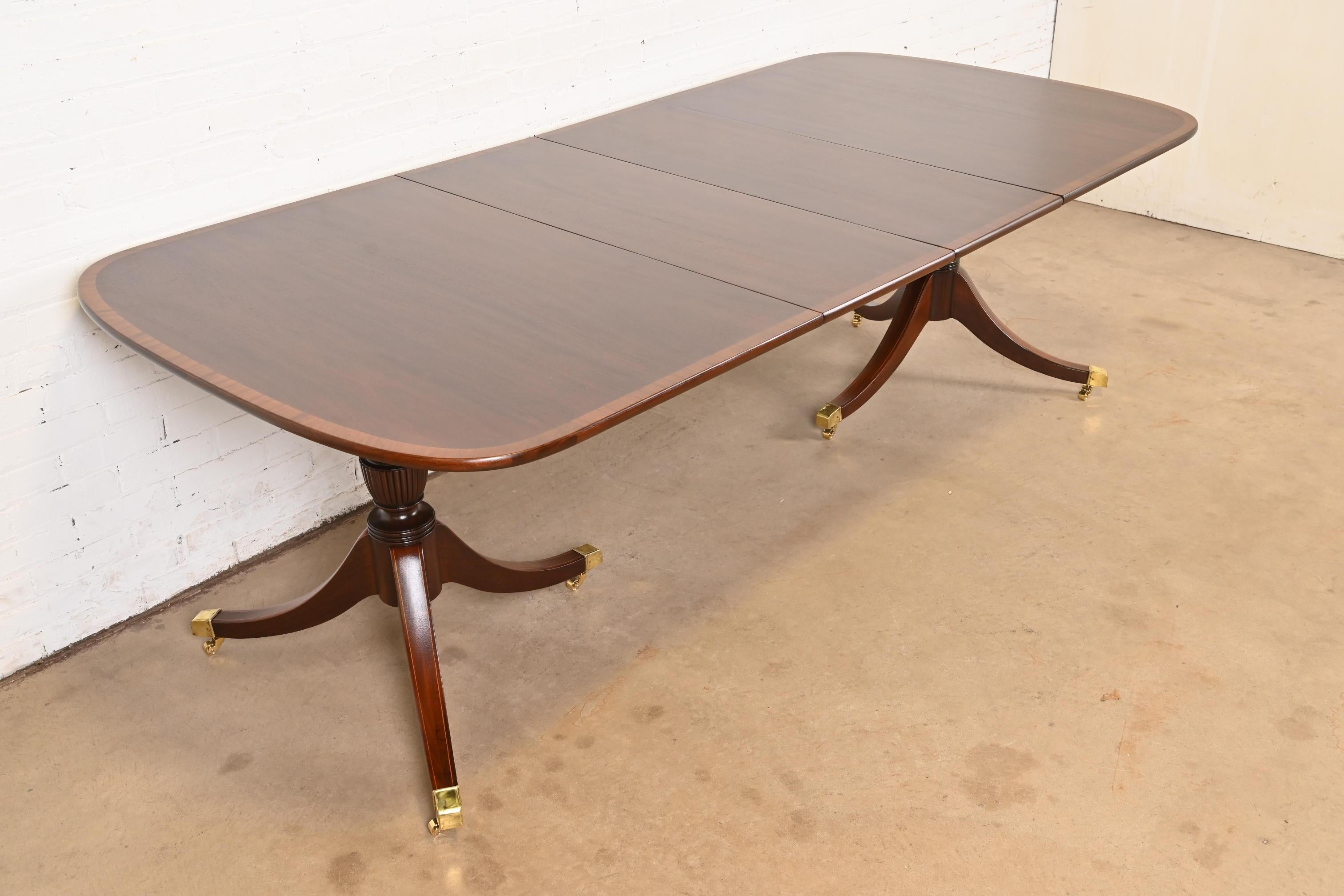 An exceptional Georgian or Regency style double pedestal extension dining table

In the manner of Baker Furniture

USA, Circa 1980s

Beautiful mahogany, with satinwood banding, carved solid mahogany pedestals, and brass-capped feet on brass