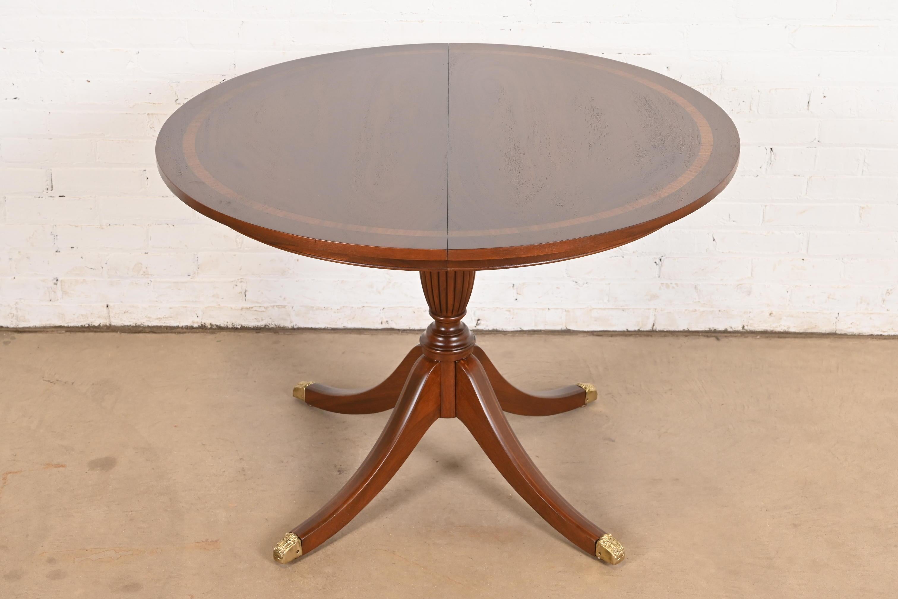 An exceptional Georgian or Regency style pedestal extension dining table

In the manner of Baker Furniture

USA, Circa 1980s

Gorgeous book-matched mahogany top, with satinwood banding, carved solid mahogany pedestal, and brass capped