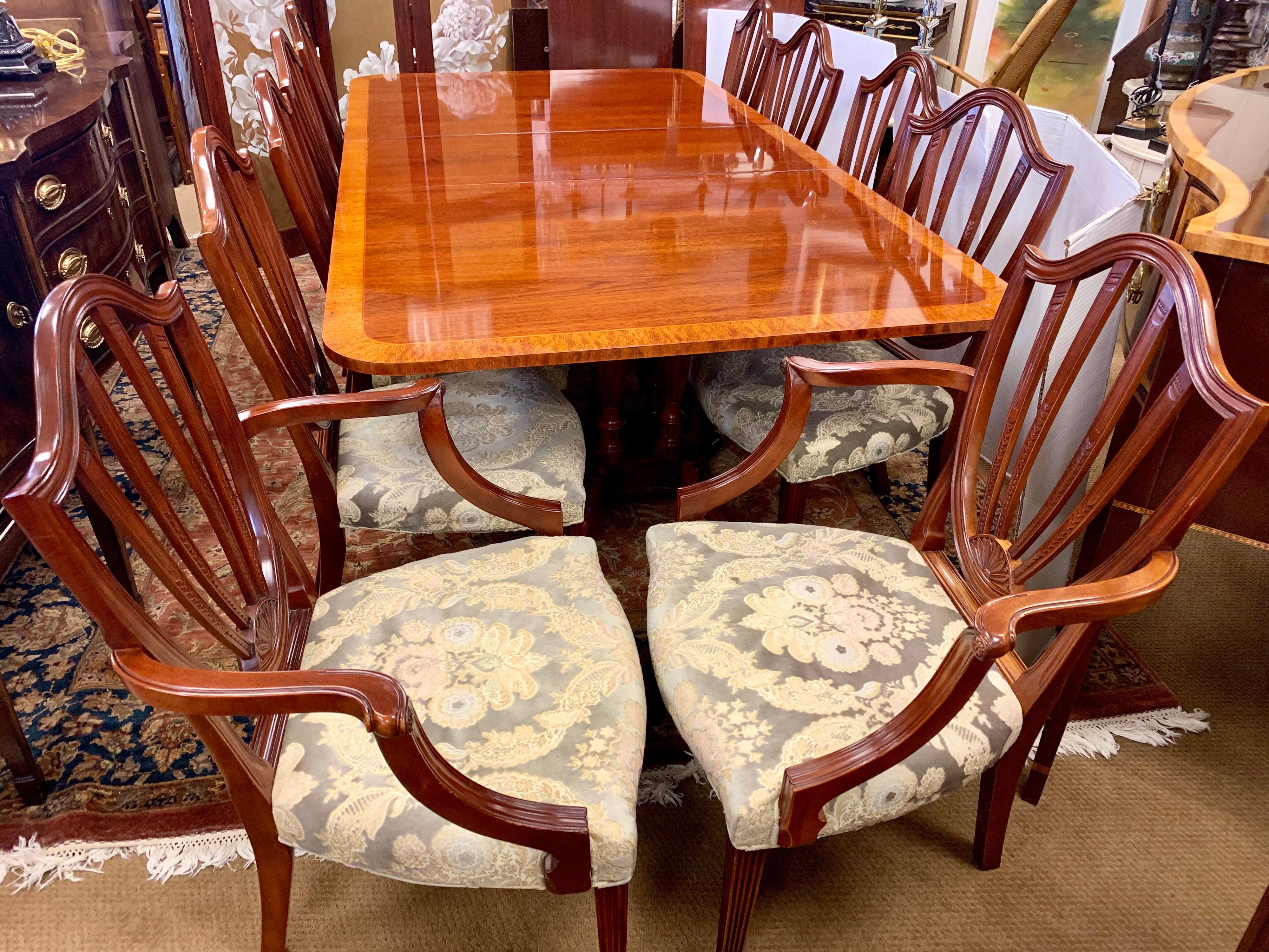Stunning Baker Furniture dining room set includes a double pedestal mahogany table with three leaves; ten shieldback chairs which include two arms and eight side chairs. Table features a banded inlay all around and brass paw feet. All Baker