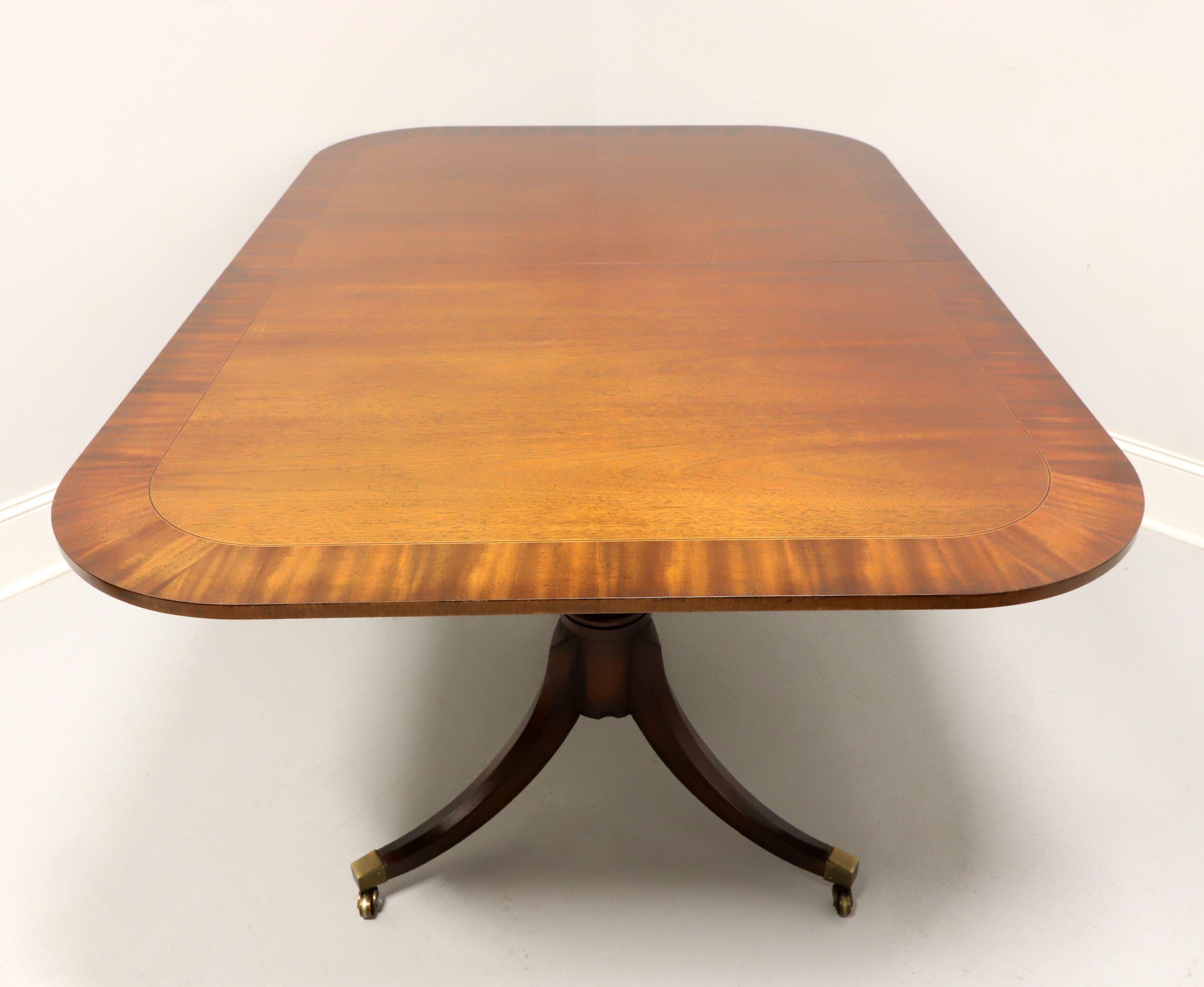 A Traditional Sheraton style dining table by Baker Furniture Company, from their Historic Charleston Collection. Mahogany, banded top with string inlay, smooth edge, carved double pedestals with three legs, brass toe caps and casters. Includes two