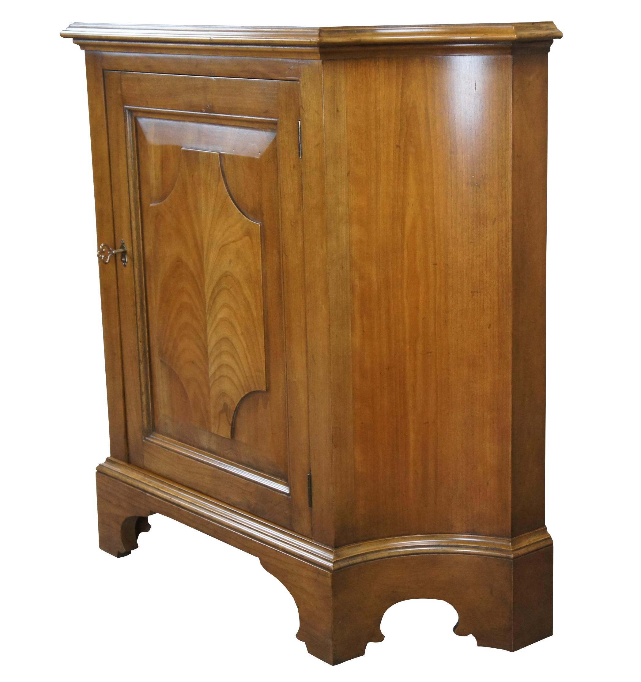 A stunning console cabinet or commode by Baker Furniture, circa 1960s. Made from Merisier (French Cherry) with a central door featuring a raised panel and elegant matchbook veneer in the design of a feather pull. The cabinet opens via skeleton key