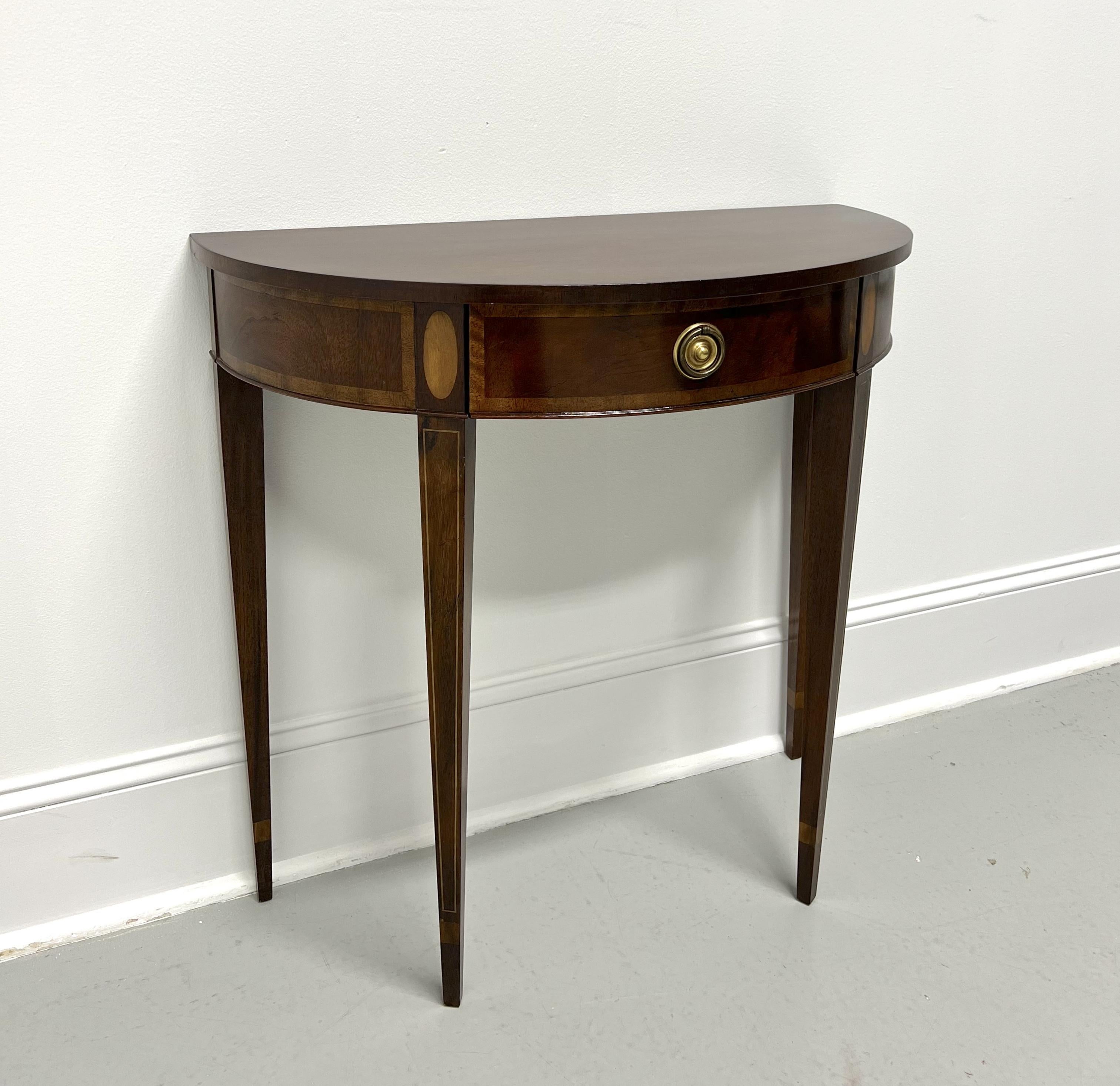 A Hepplewhite style demilune console table by Baker Furniture. Mahogany with brass hardware, banded & inlaid apron, banded drawer front, and tapered straight legs with string inlays. Features one drawer of dovetail construction. Made in the USA, in
