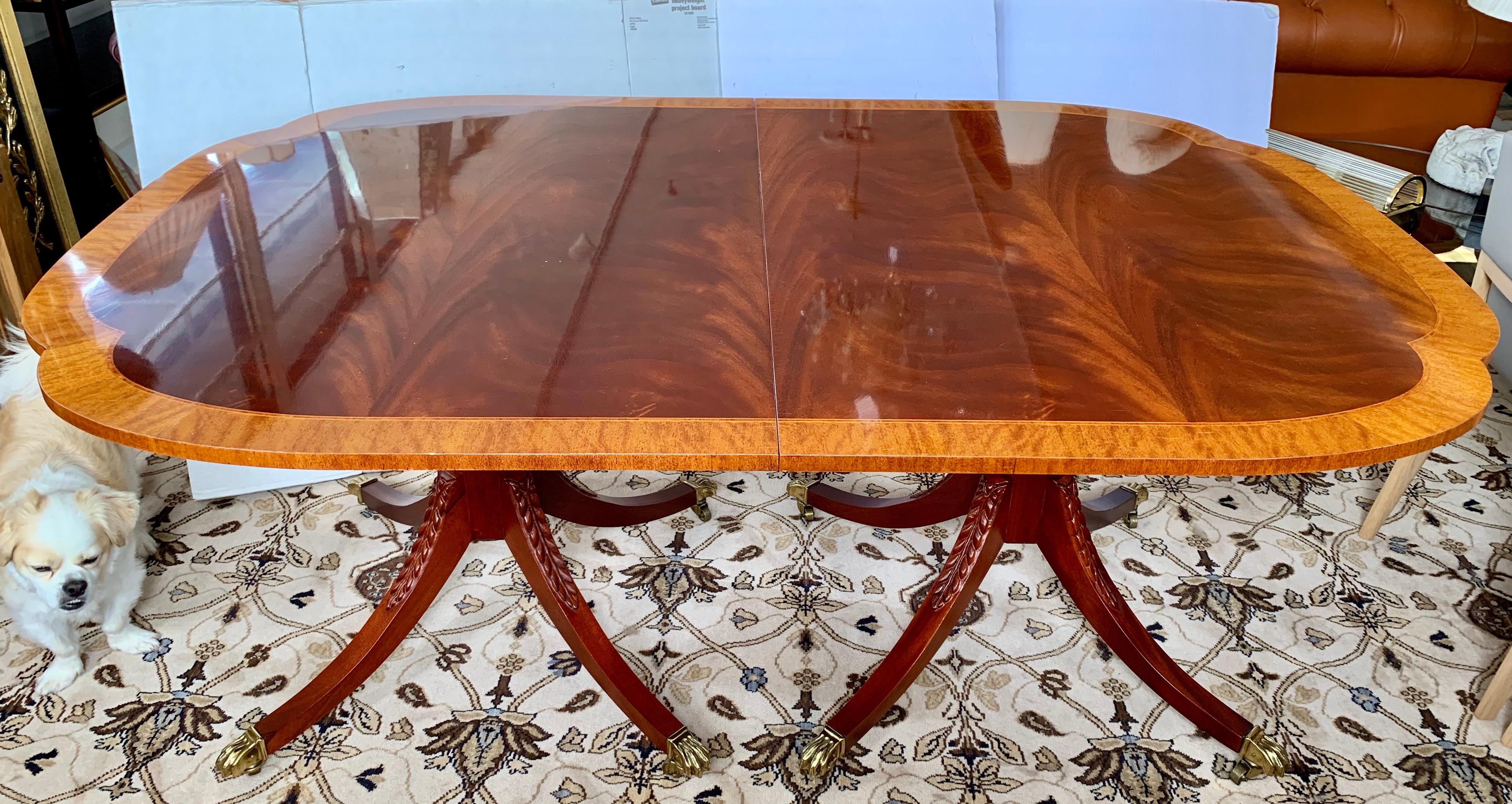 Fabulous quality book-matched flame mahogany double pedestal, three leaves, scalloped edge dining table. Features satinwood banding (inlay) and splay-legged pedestals with carved acanthus leaf detail on cast brass lion's paw feet with castors.