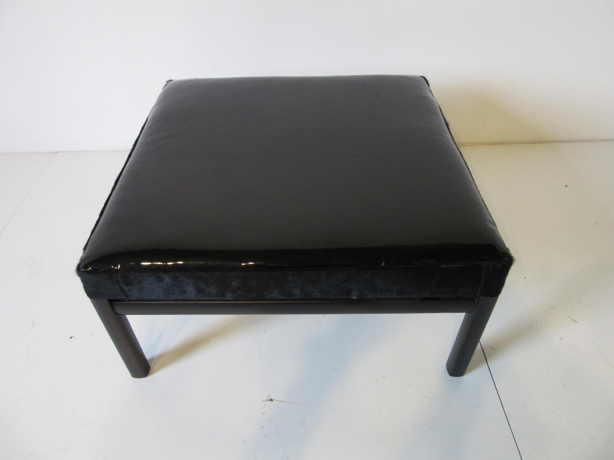 A ebony toned wood framed ottoman with brass details, upholstered in black patent leather, sides trimmed in black pony hide. A stunning twist on a great design by Baker and Micheal Taylor.