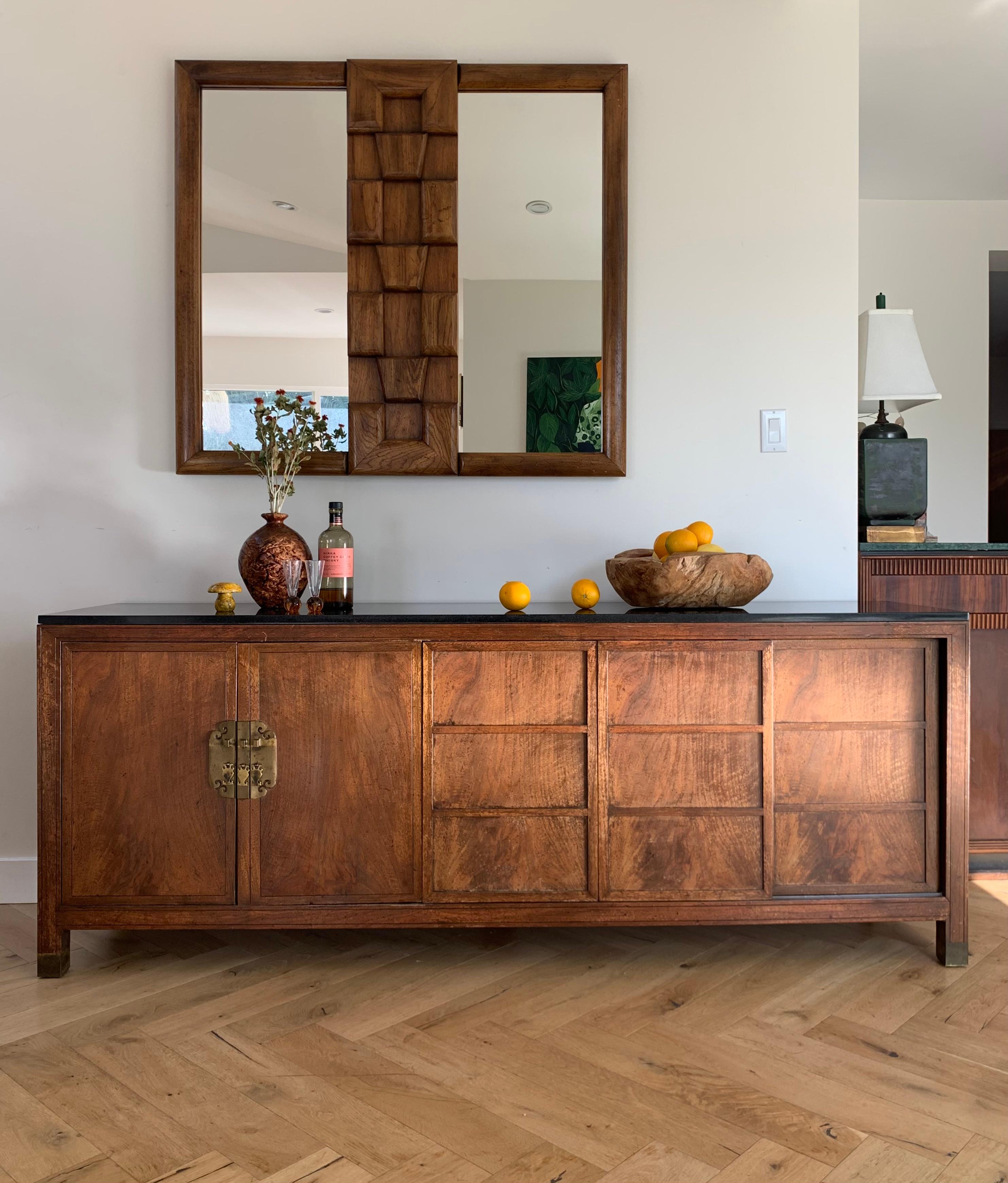 A stately mahogany wood credenza / sideboard / buffet with brass accents and granite top by Baker Furniture Company, 1950s. At once rustic and regal, this piece fuses mid century modern sensibilities with far-east oriental detailing. The right side