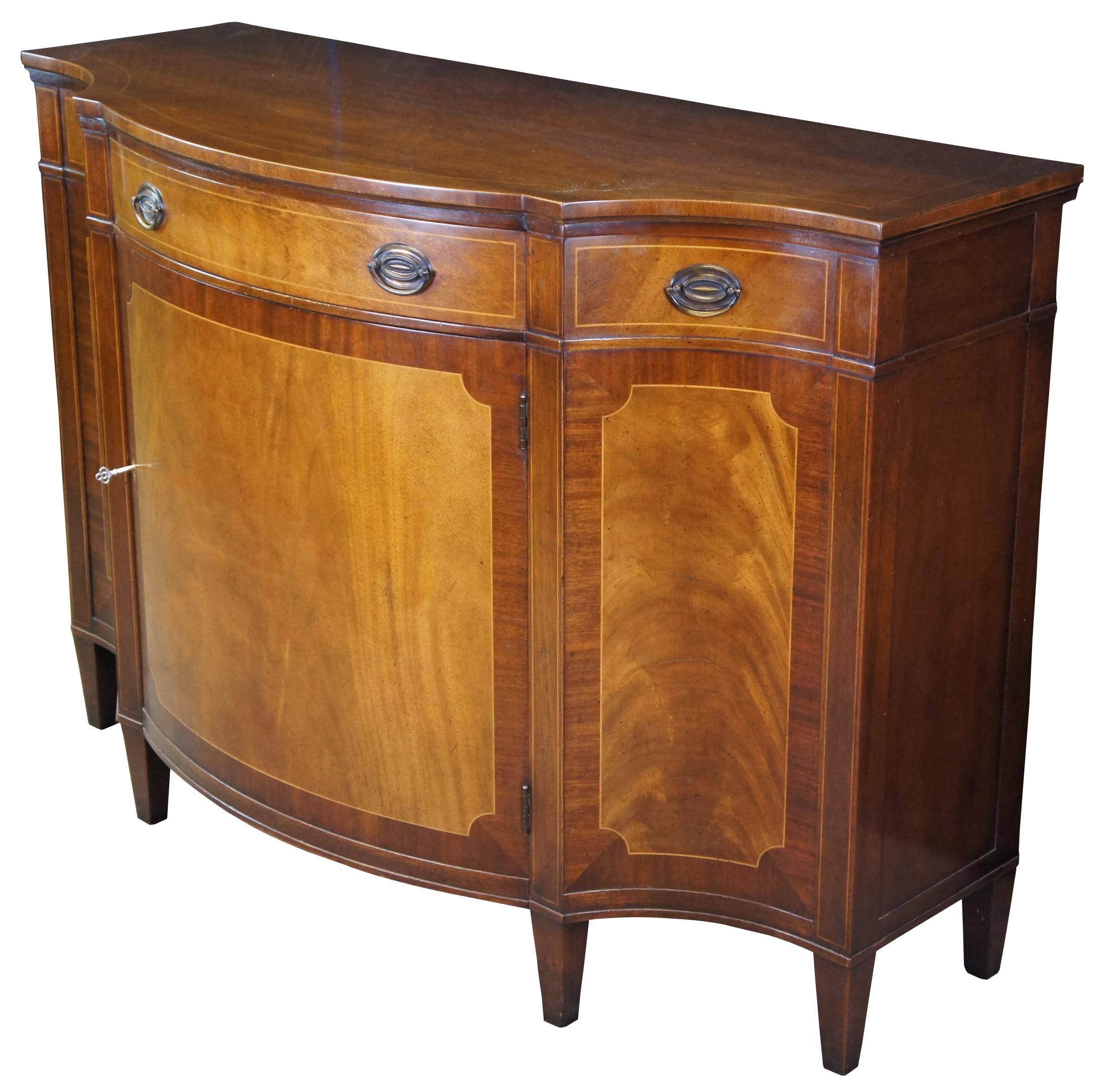 Vintage Baker Furniture Sheraton style server or console cabinet. Made from Mahogany with banded crotch or flame veneered front. Features a bowfront with central drawer over large lower storage cabinet. The cabinet is supported by square tapered