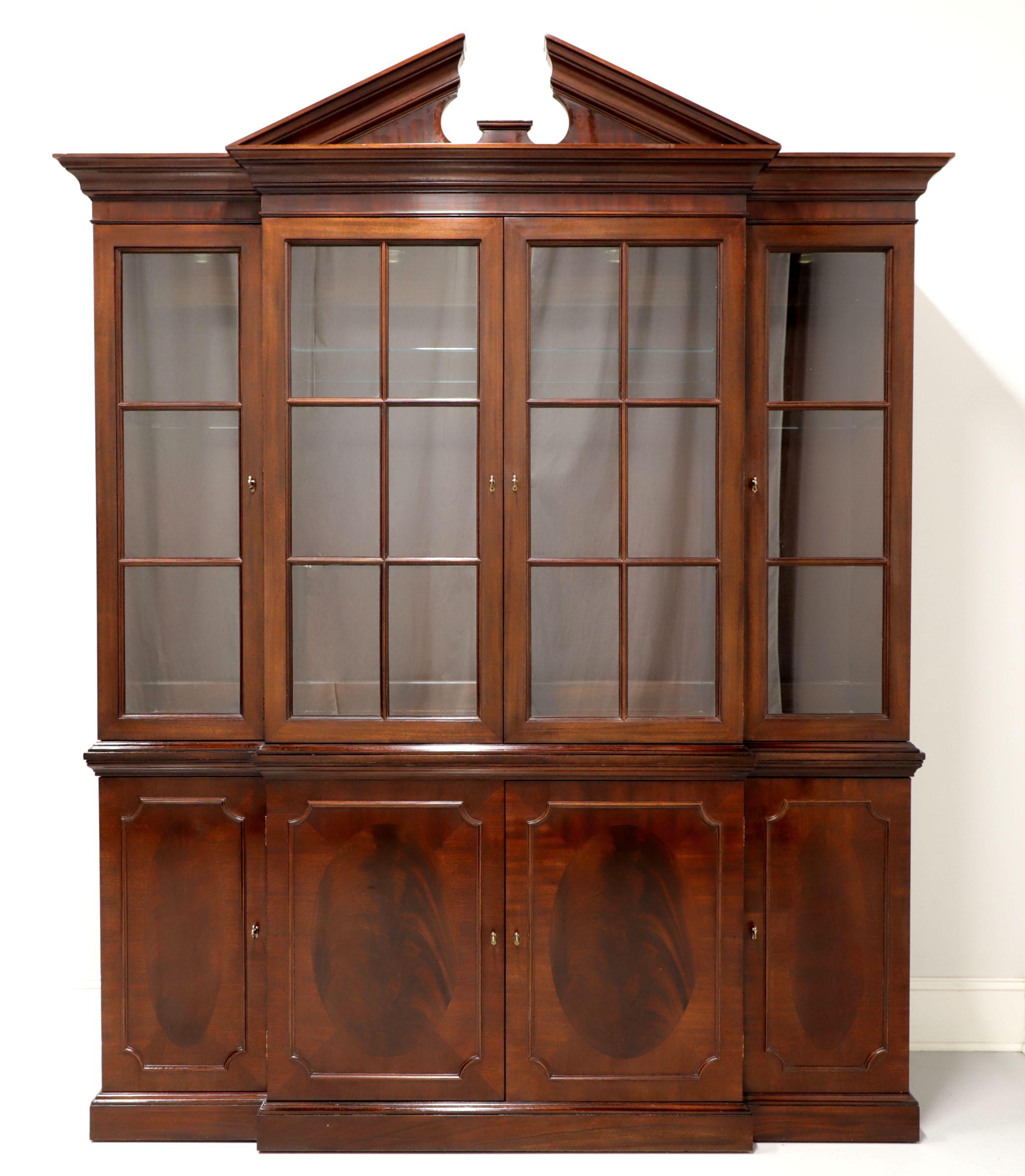 A Traditional style breakfront china cabinet by Baker Furniture. Mahogany with Inlaid flame mahogany, brass hardware, crown moulding with pediment to top and paned glass doors. Upper lighted cabinet features three separate compartments with two