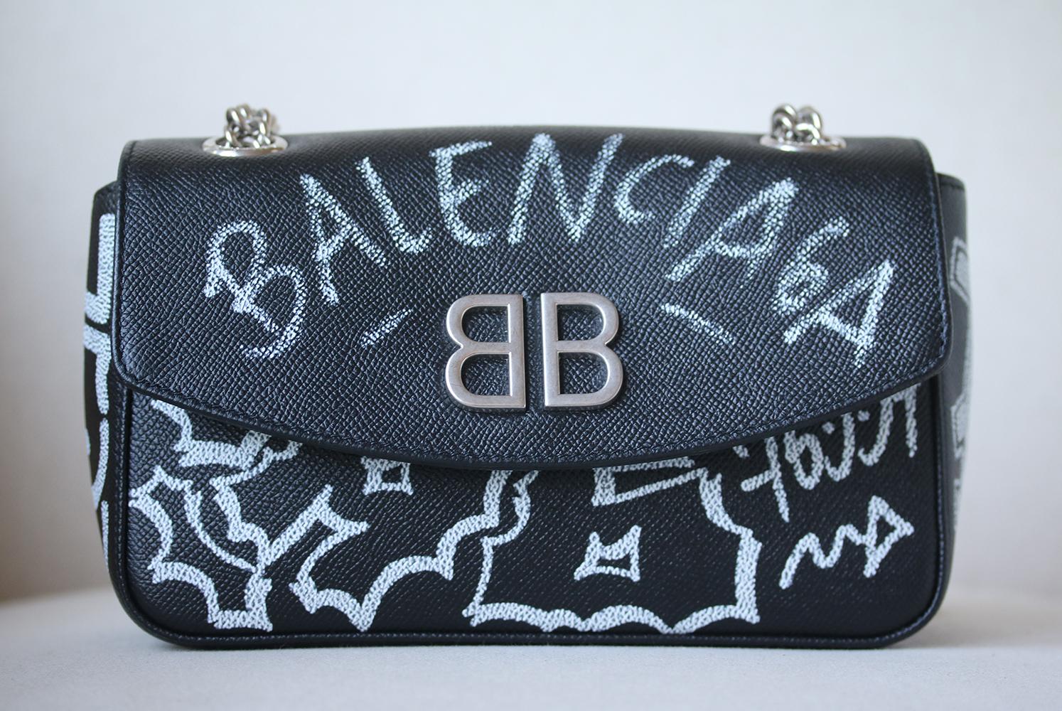Balenciaga’s BB cross-body bag gets a rebellious revamp for Pre-AW18. This black leather iteration is suspended from a silver-tone metal chain and leather cross-body strap, and features irreverent graffiti phrases painted across the exterior,