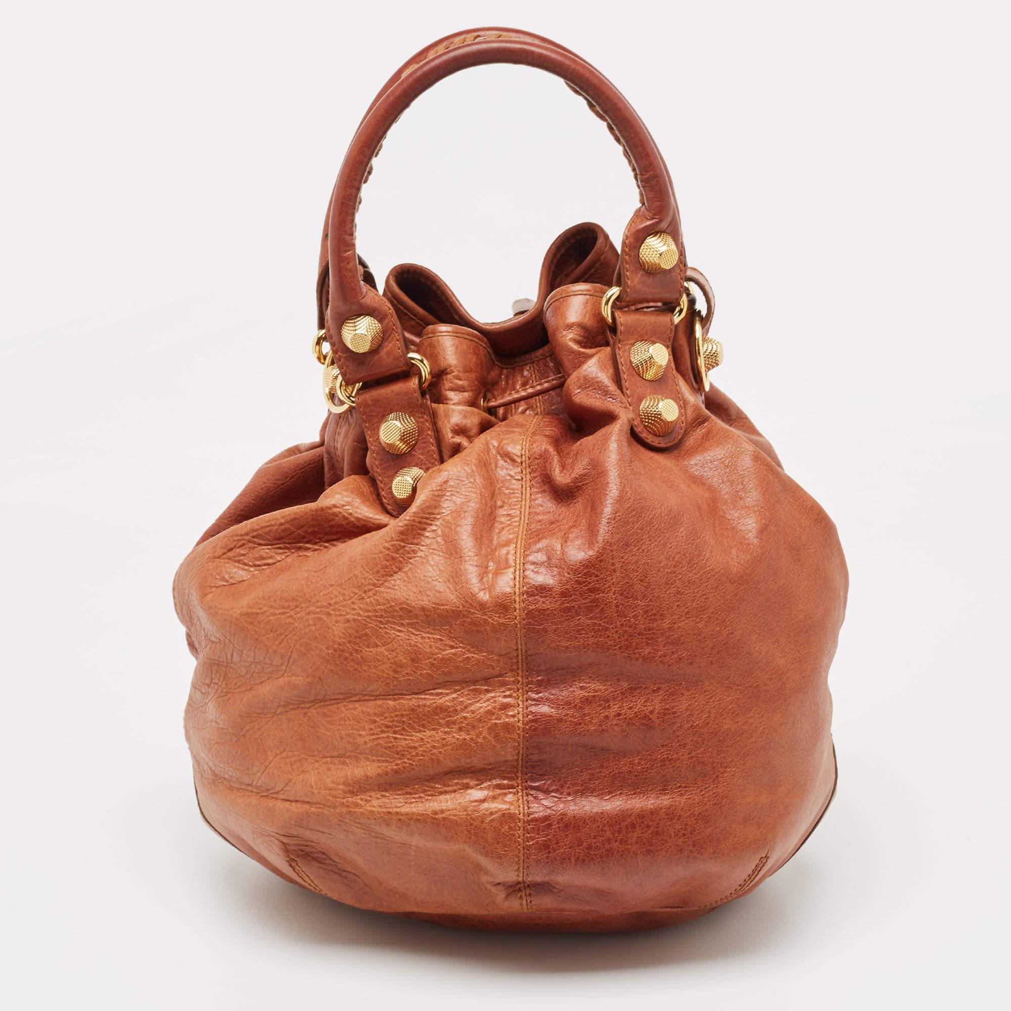 Ce sac 'PomPon' de Balenciaga deviendra le complément idéal de votre garde-robe de jour ! Il est réalisé en cuir marron et rehaussé de clous en métal doré et d'autres détails matériels. L'extérieur est agrémenté de poignées roulées, d'une poche