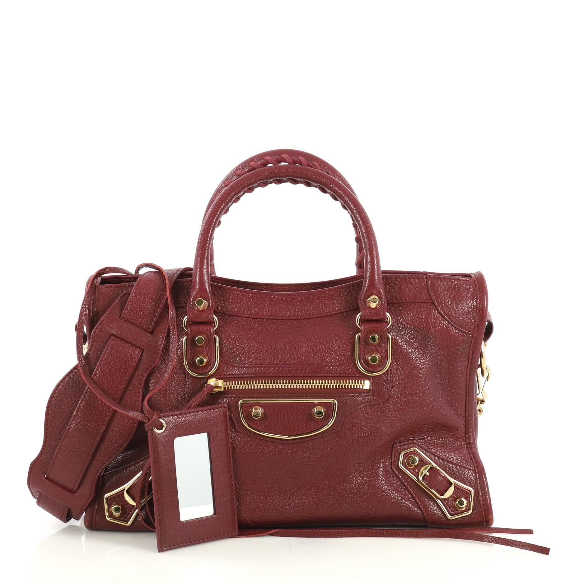 This Balenciaga City Classic Metallic Edge Bag Leather Small crafted from red leather, features dual braided woven handles, studs and buckle details, front zip pocket, and gold-tone hardware. Its top zip closure opens to a black fabric interior with