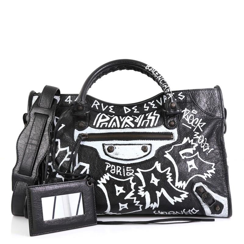 This Balenciaga City Graffiti Classic Studs Bag Leather Medium, crafted from black printed leather, features dual braided woven handles, multicolor printed graffiti writing, front zip pocket, stud and buckle detailing, and bronze-tone hardware. Its