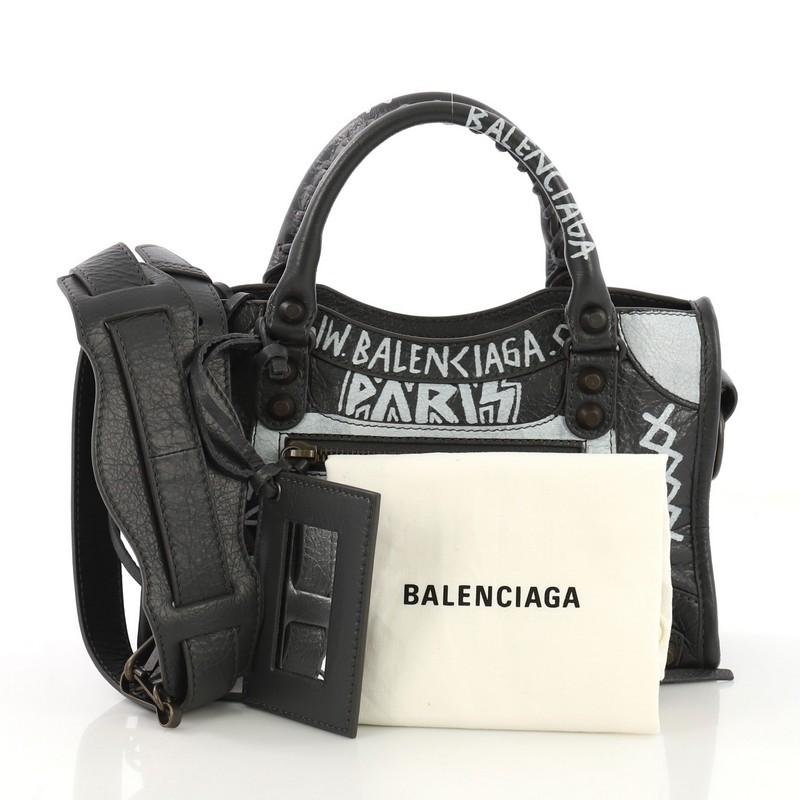 This Balenciaga City Graffiti Classic Studs Bag Leather Mini, crafted from gray leather, features dual braided woven handles, front zip pocket, printed graffiti writing, and brass-tone hardware. Its top zip closure opens to a black fabric interior
