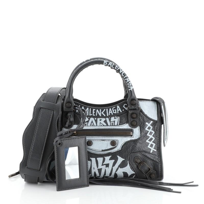 This Balenciaga City Graffiti Classic Studs Bag Leather Mini, crafted from gray leather, features dual braided woven handles, front zip pocket, printed graffiti writing, and antique brass-tone hardware. Its top zip closure opens to a black fabric