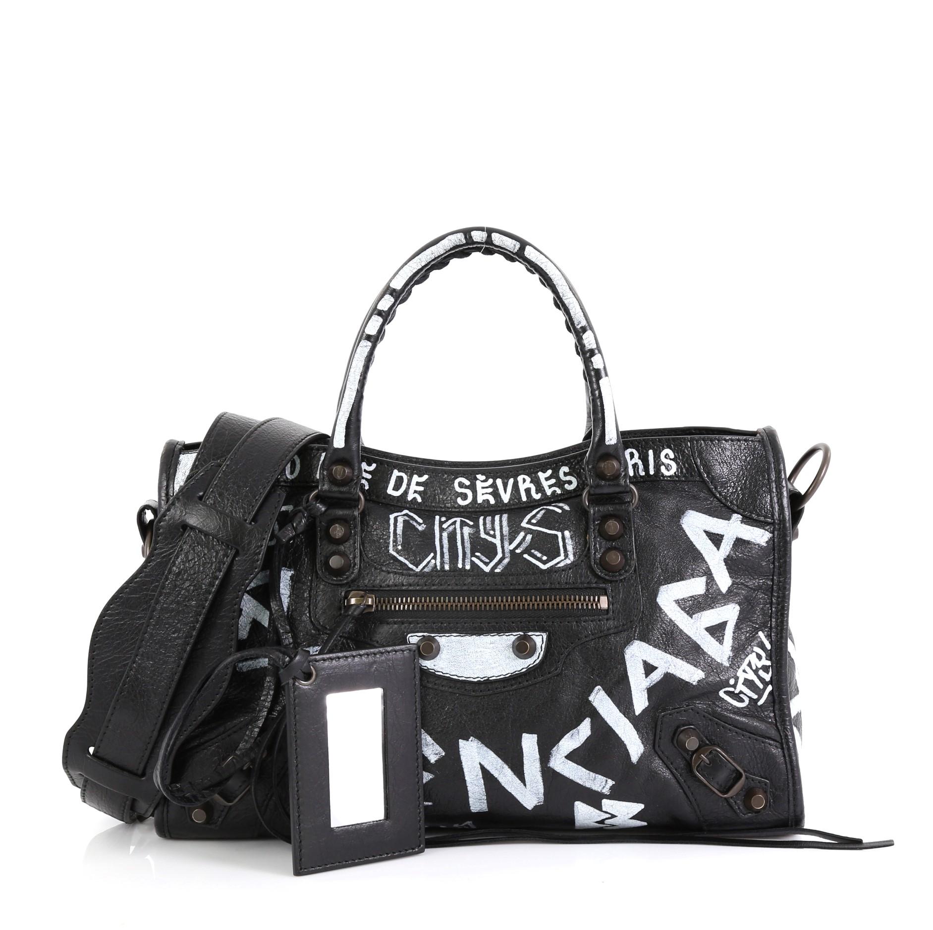 This Balenciaga City Graffiti Classic Studs Bag Leather Small, crafted from black leather, features dual braided woven handles, front zip pocket, printed graffiti writing, and brass-tone hardware. Its top zip closure opens to a black fabric interior