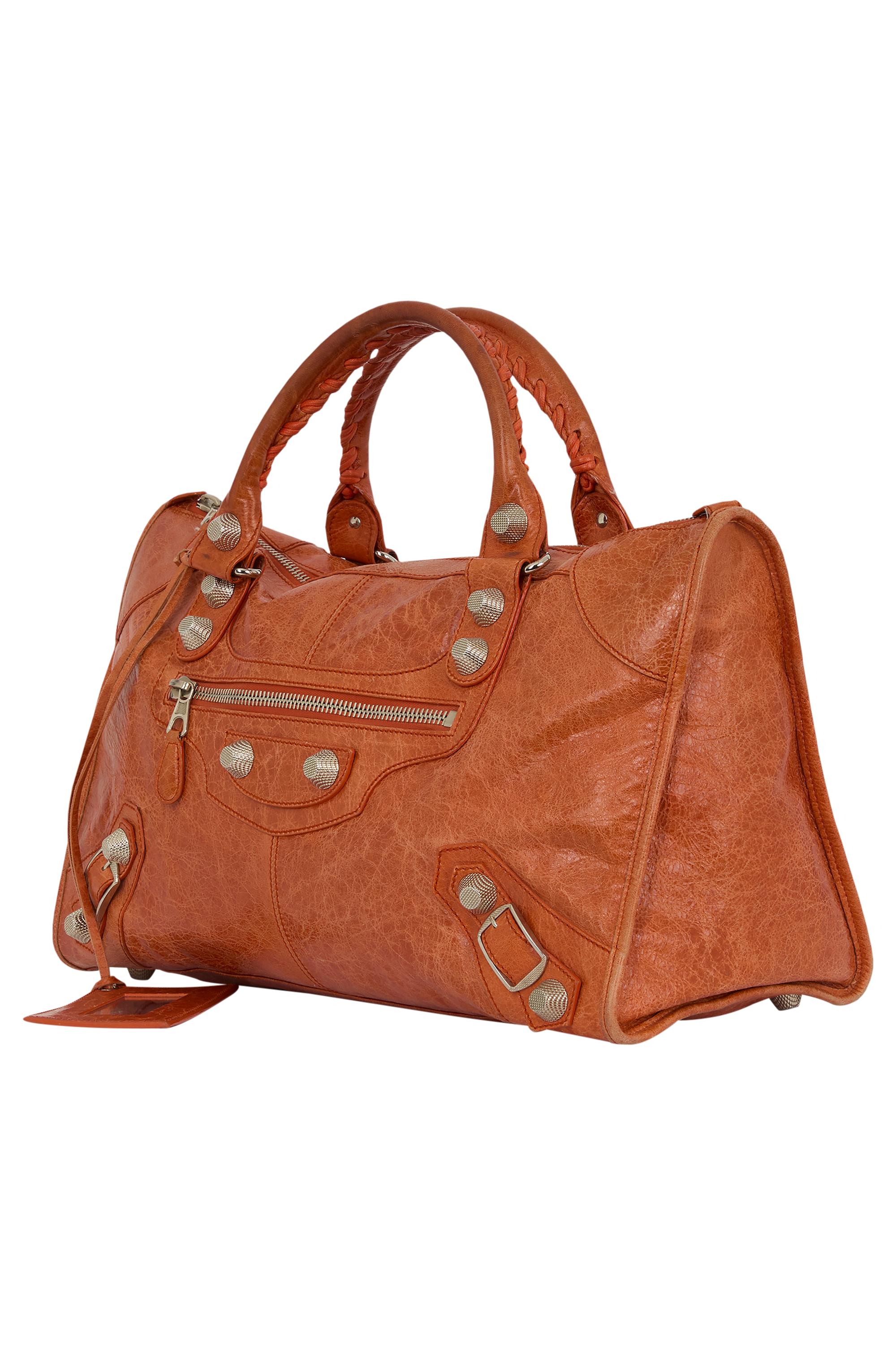 Pièce emblématique et intemporelle de Balenciaga, le sac City Classic incarne le cool sans effort. Confectionné en cuir souple orange avec des accessoires argentés, il présente une poche zippée sur le devant, une fermeture zippée sur le dessus avec