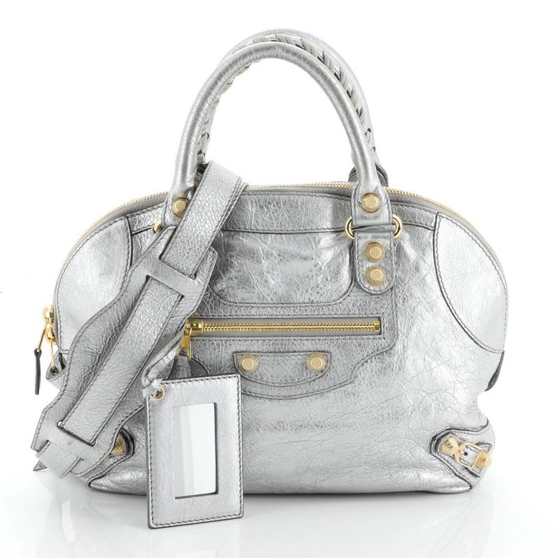 This Balenciaga Convertible Bowling Bag Classic Studs Leather Mini crafted in grey leather, features dual braided woven handles, front zip pocket, iconic classic studs and buckle details, leather fringes zip pull, and gold-tone hardware. Its top zip