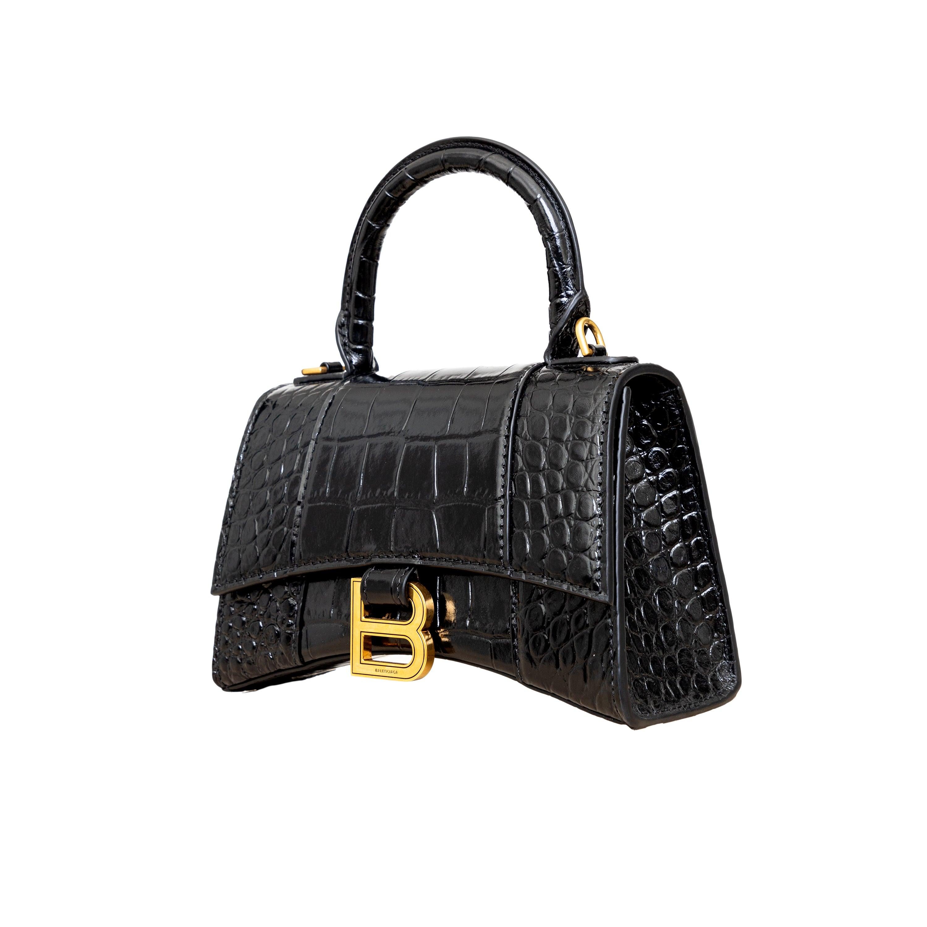 Crafted from black, shiny crocodile embossed calfskin, this Balenciaga handbag in XS format features the iconic curvilinear base, one top handle, and a studded magnet closure for a chic and modern touch. The aged-gold B logo hardware and nappa