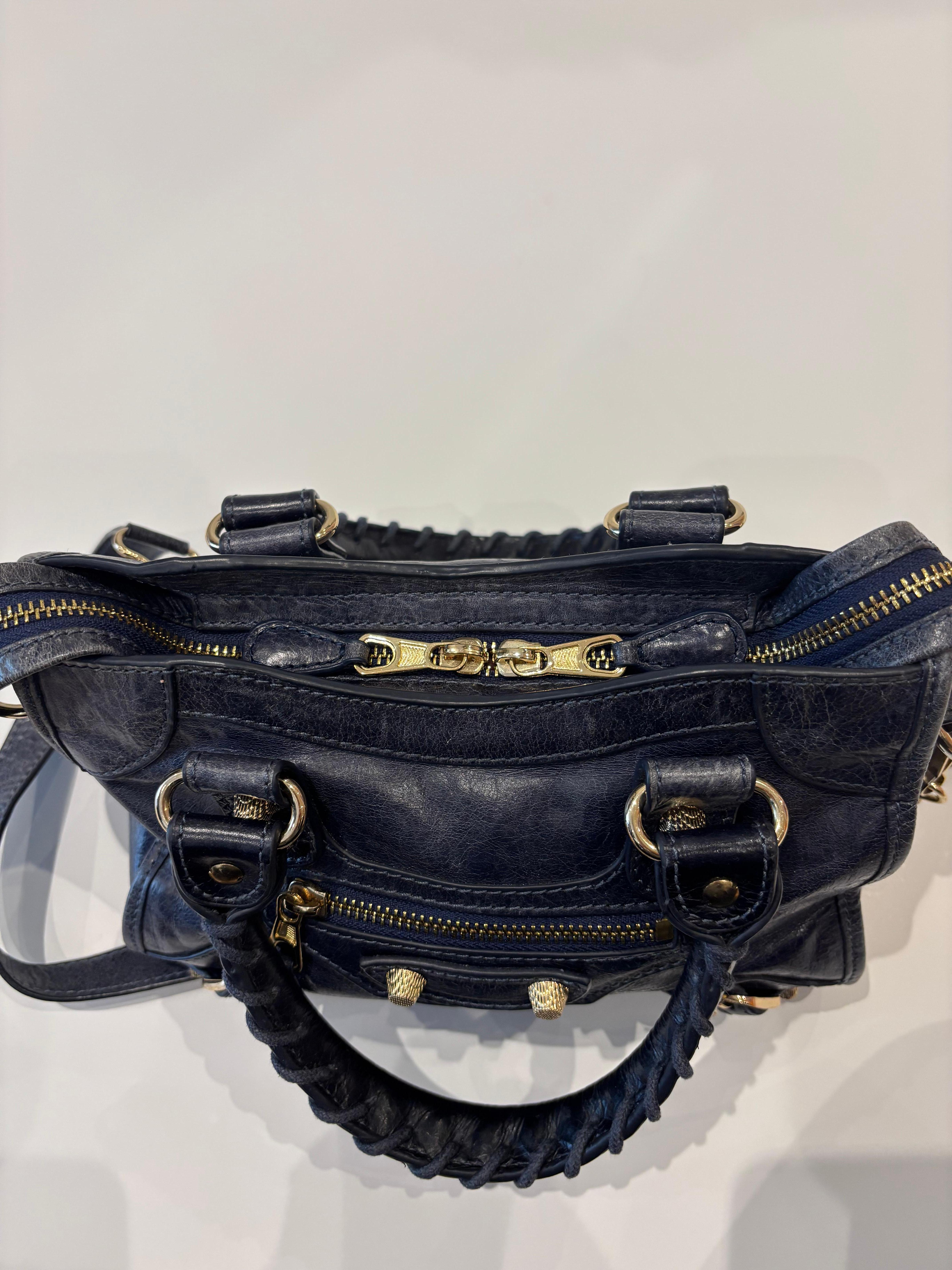 This is the Balenciaga Classic City Mini leather two way bag, with top braided handles and a removeable crossbody strap.
The color is a dark blue aged leather with gold hardware. The hardware includes lovely stylized grommets.
There is a little wear