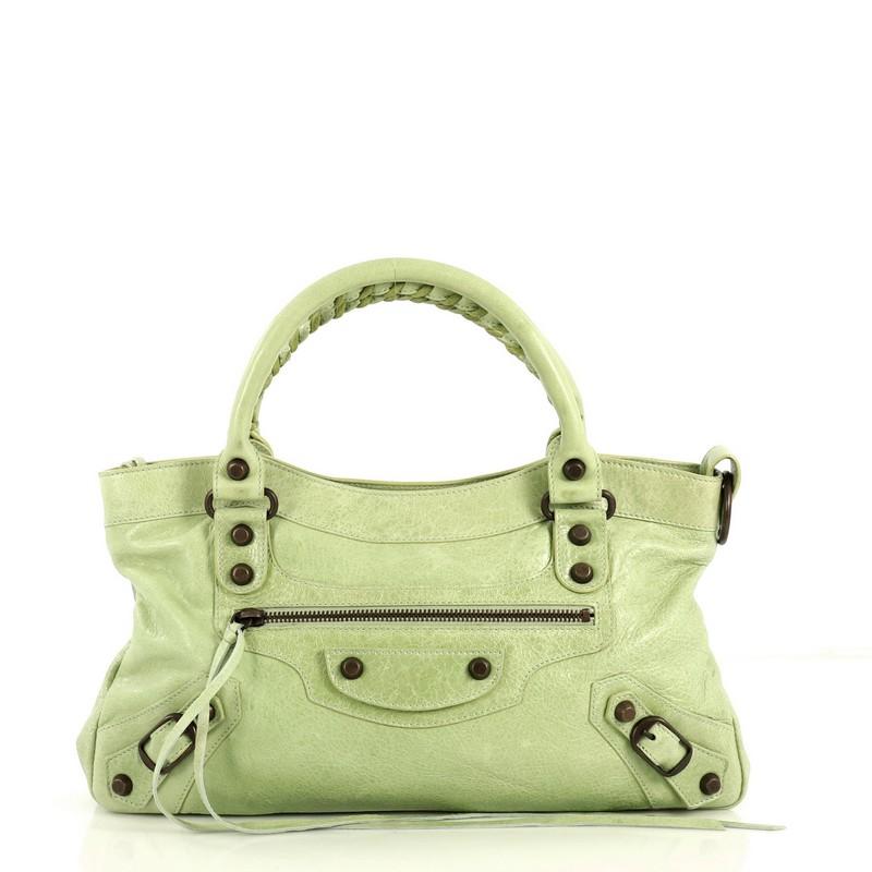 This Balenciaga First Classic Studs Bag Leather, crafted from light green leather, features braided woven handles, exterior front zip pocket, classic studs and buckle details, and brass-tone hardware. Its top zip closure opens to a black fabric