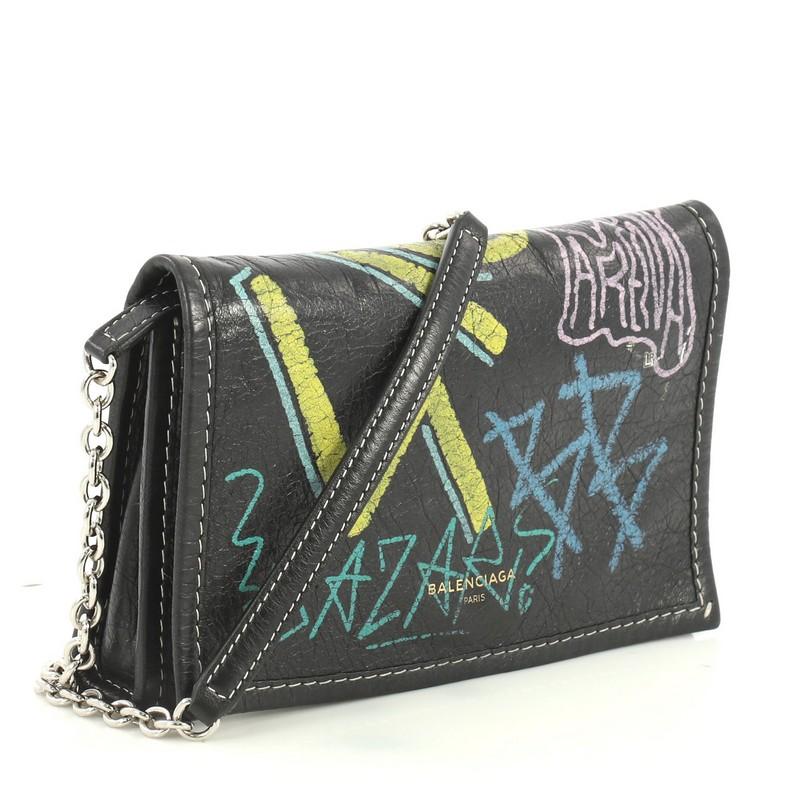 This Balenciaga Graffiti Wallet on Chain Leather, crafted from black printed leather, features chain link strap, multicolor printed graffiti writing, and silver-tone hardware. Its magnetic snap closure opens to a black leather and fabric interior