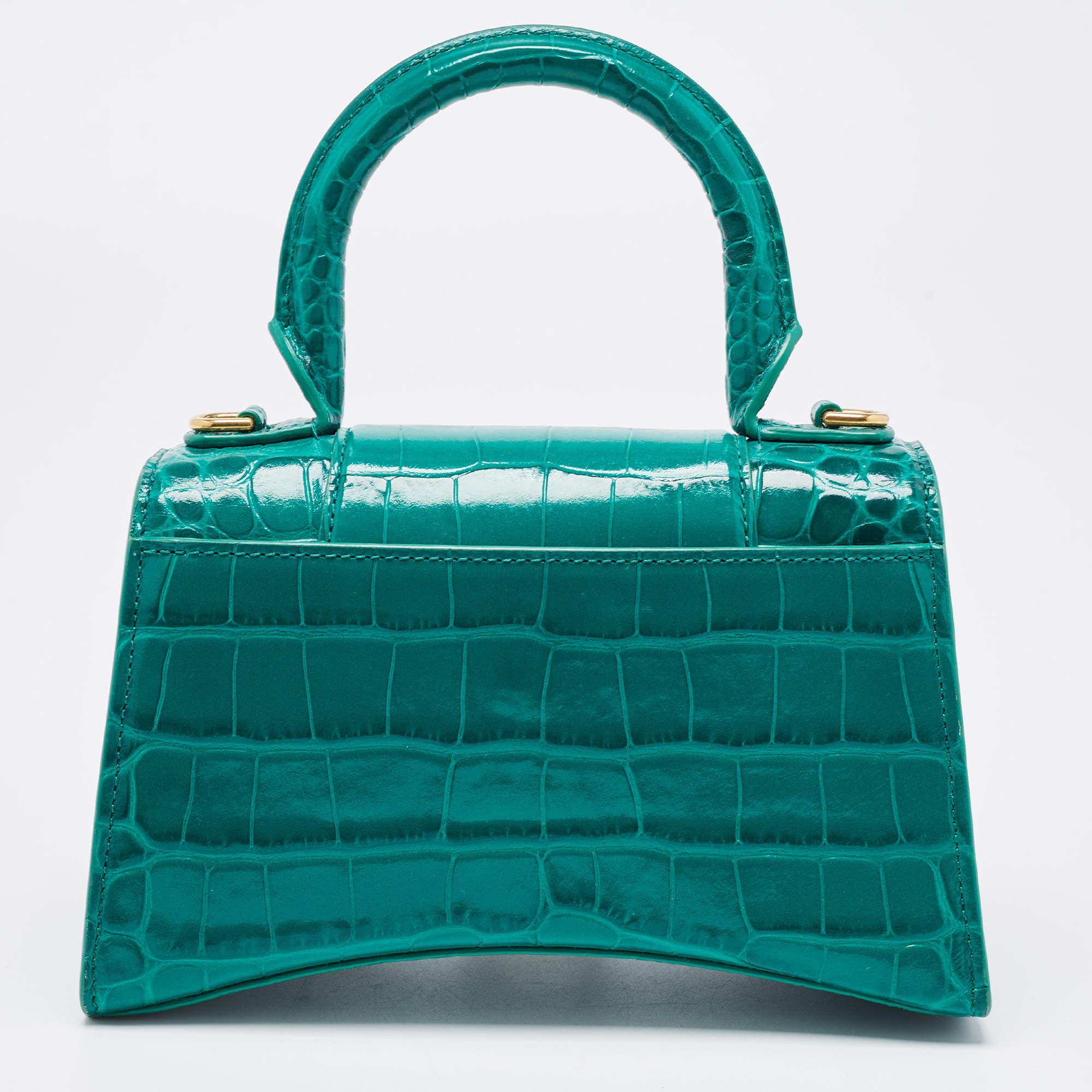 The Hourglass features an croc-embossed leather body, a rolled top handle, a detachable flat leather shoulder strap, a front flap with magnetic snap closure, and a spacious interior.

