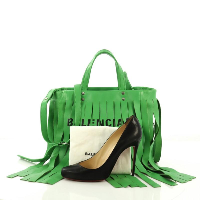 This Balenciaga Laundry Cabas Tote Fringe Leather XS, crafted in green fringe leather, features dual flat top handles and silver-tone hardware. It opens to a black fabric interior with zip and slip pockets. **Note: Shoe photographed is used as a