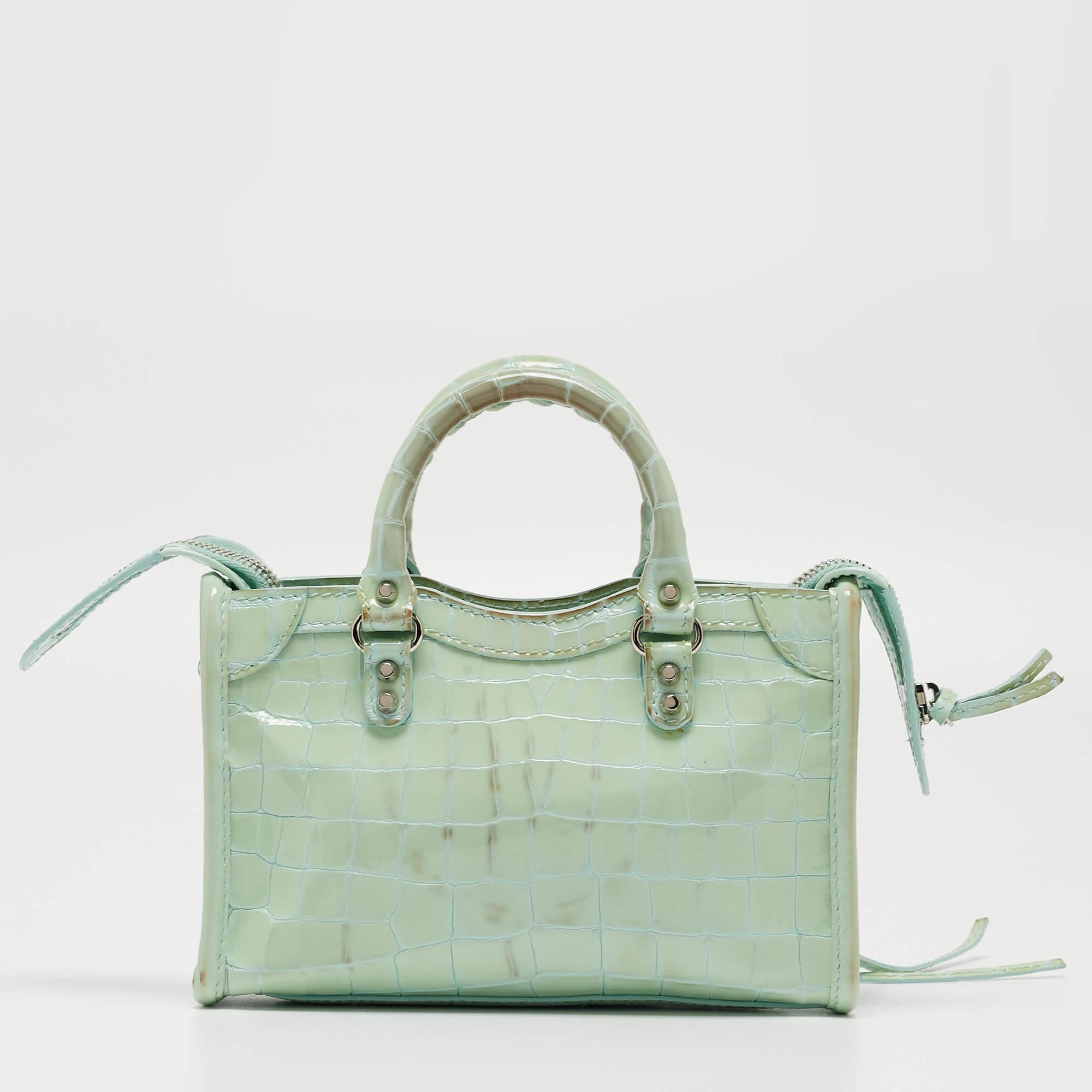 The Balenciaga Nano Classic City Tote exudes luxury with its vibrant mint green hue and textured croc-embossed pattern. Crafted from high-quality patent leather, this compact tote features the iconic City silhouette, making it a stylish and
