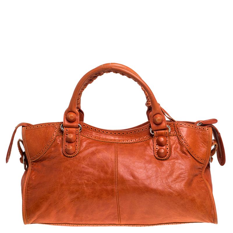 This Part Time tote from Balenciaga is perfect for all your outings. This orange leather bag is unique in its silhouette and features an interplay of large studs and buckles. With a creative edge, the bag has a front zip pocket, round leather