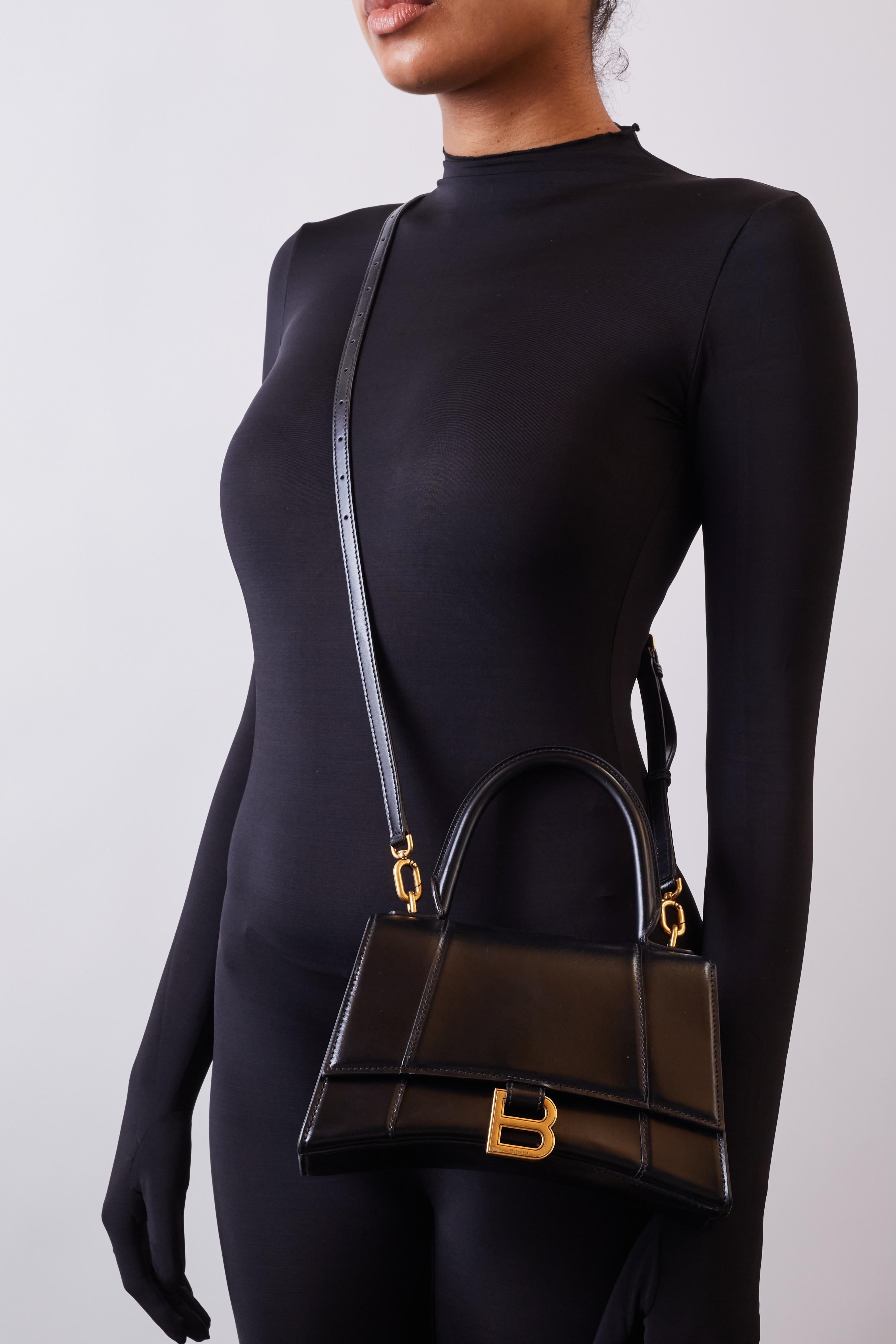 This petite handbag is made of smooth calfskin leather in black. The bag features a rolled leather top handle, the signature Balenciaga B charm at snap closure, gold hardware, and slip pocket at the back. The front flap opens to a black leather