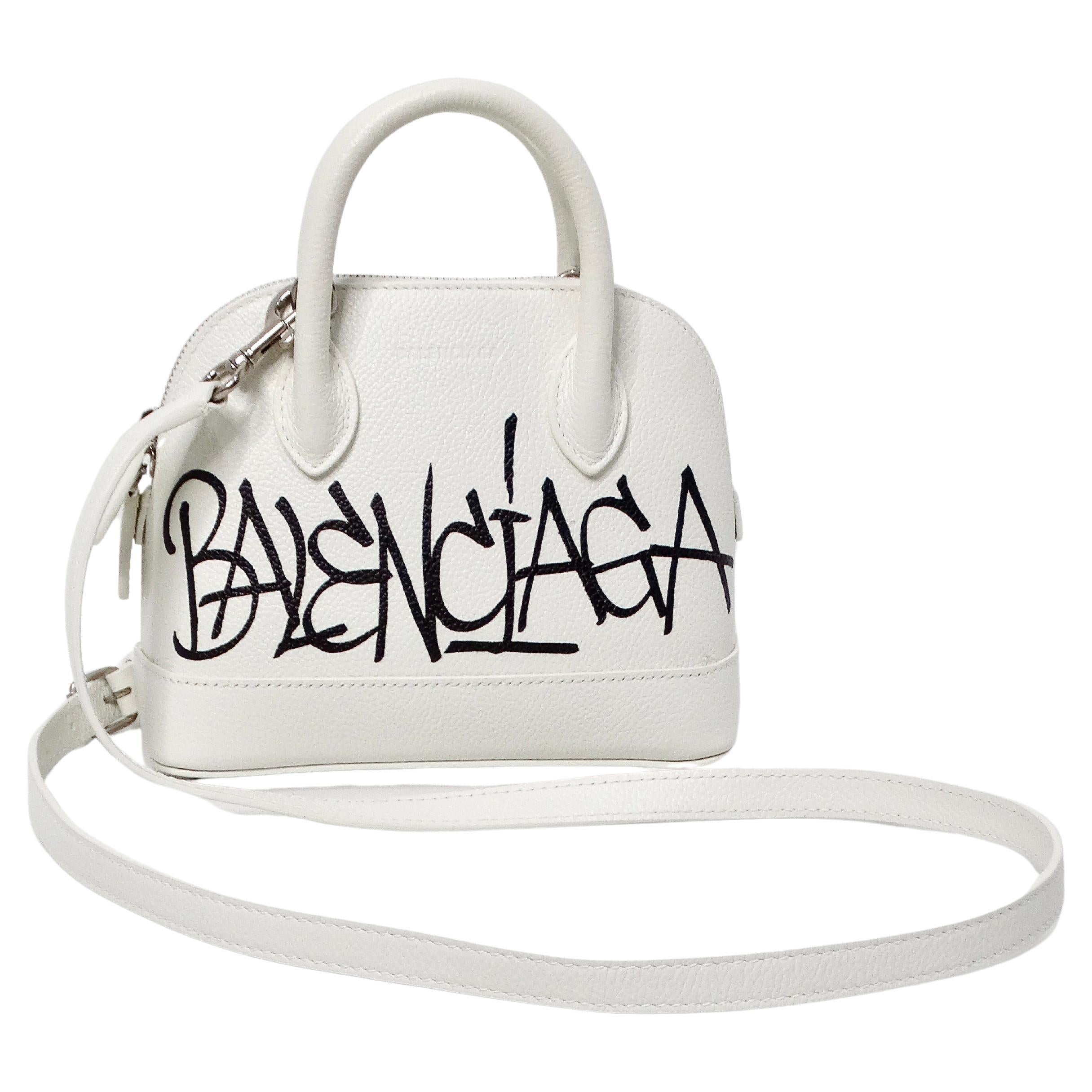 The Balenciaga White Graffiti Logo Print Ville Leather XXS Mini Top Handle Bag is a striking and contemporary accessory that effortlessly combines edgy street style with high-fashion sophistication. Crafted from white leather, this mini bag features