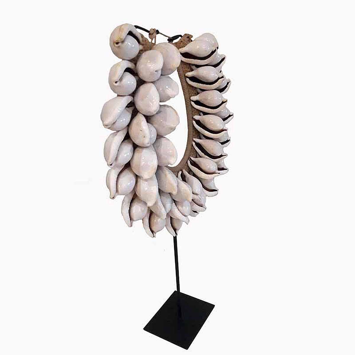 A decorative necklace from Bali, Indonesia, on a black metal stand. The large, white cowry shells are delicately attached to a handwoven cotton / raffia base. Mounted on a black metal stand.