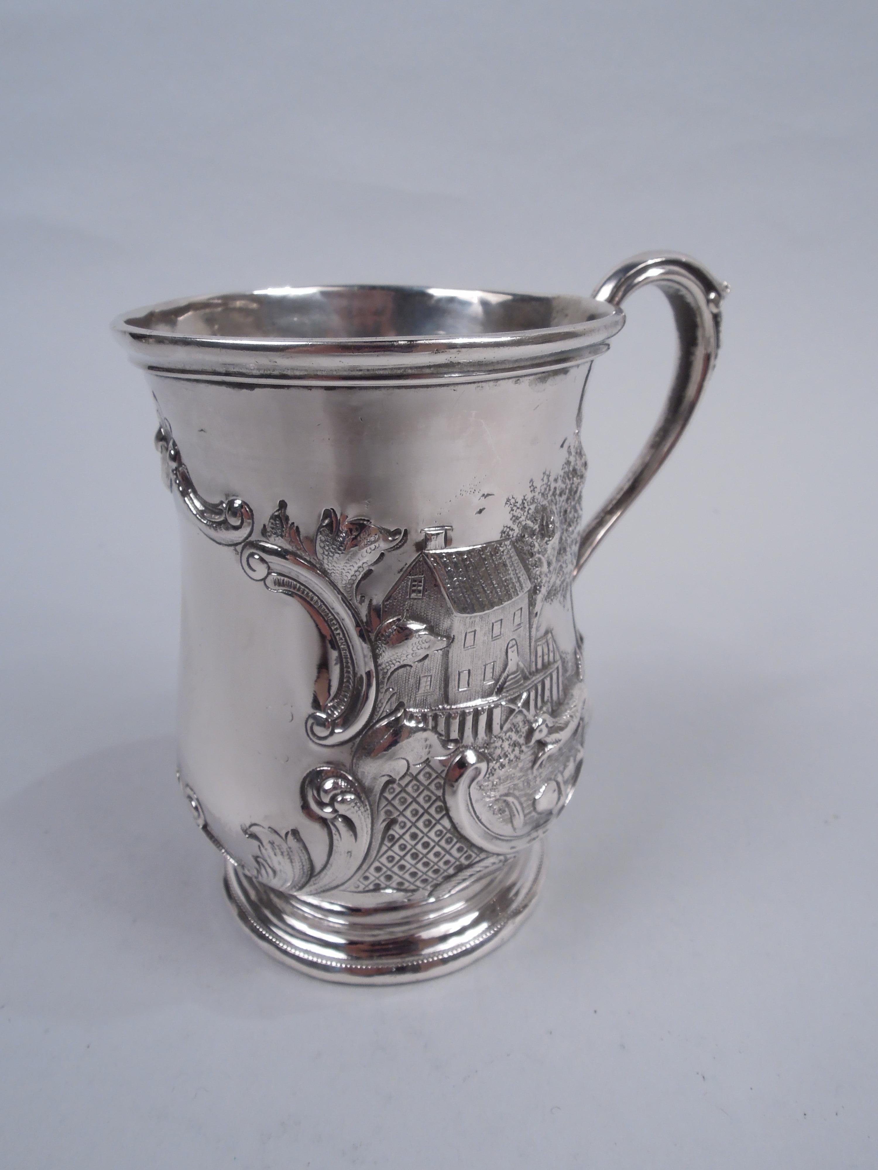 Classical coin silver christening mug, ca 1850. Baluster bowl with high-looping leaf-capped s-scroll handle and round stepped foot. Large scrolled frame (vacant) surrounded by rural idyll with house and barn set in bosky landscape. In foreground a