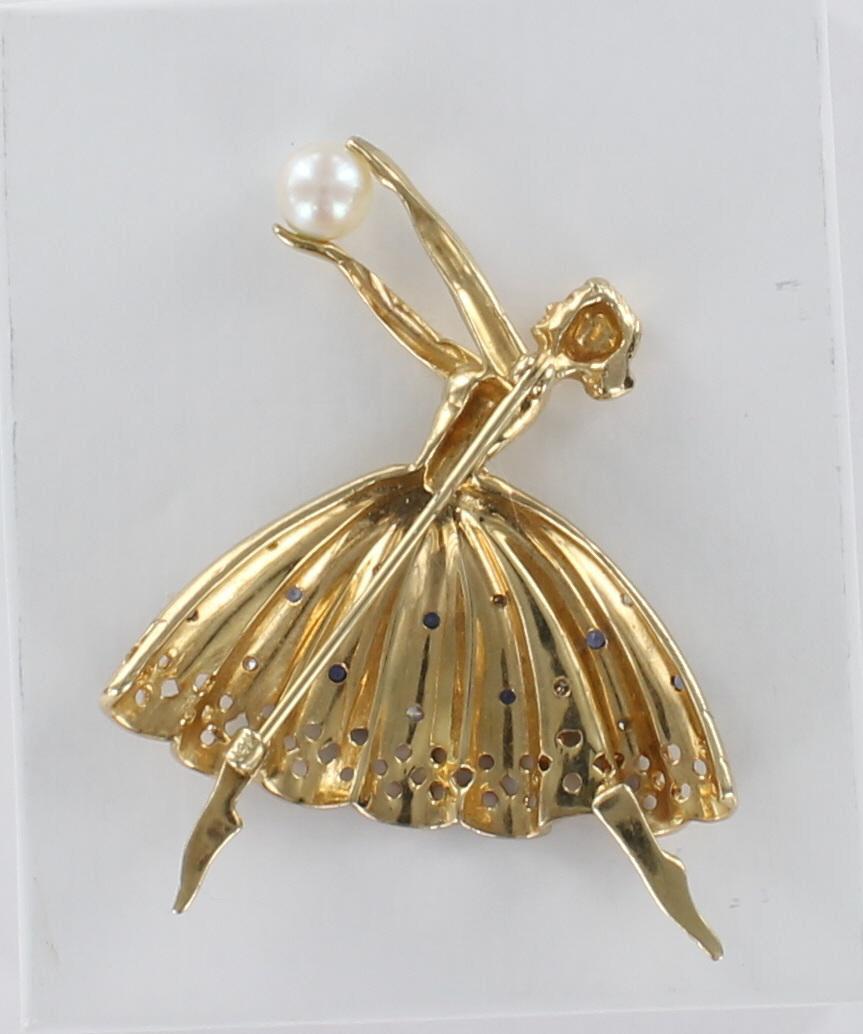 This 14 karat yellow gold pin is grand and graceful. As the ballerina is dancing, her arms are outstretched and holding a lovely pearl. Her skirt is crafted to have folds in it and is set with diamonds and sapphires. There is a row of openwork