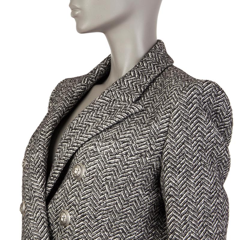 Balmain herringbone signature double-breasted blazer in black, grey, and off-white cotton (60%) and wool (40%). With peak collar, decorative lion buttons in silver metal, chest pockets, two flap pockets on the sides, and buttoned cuffs. Closes with