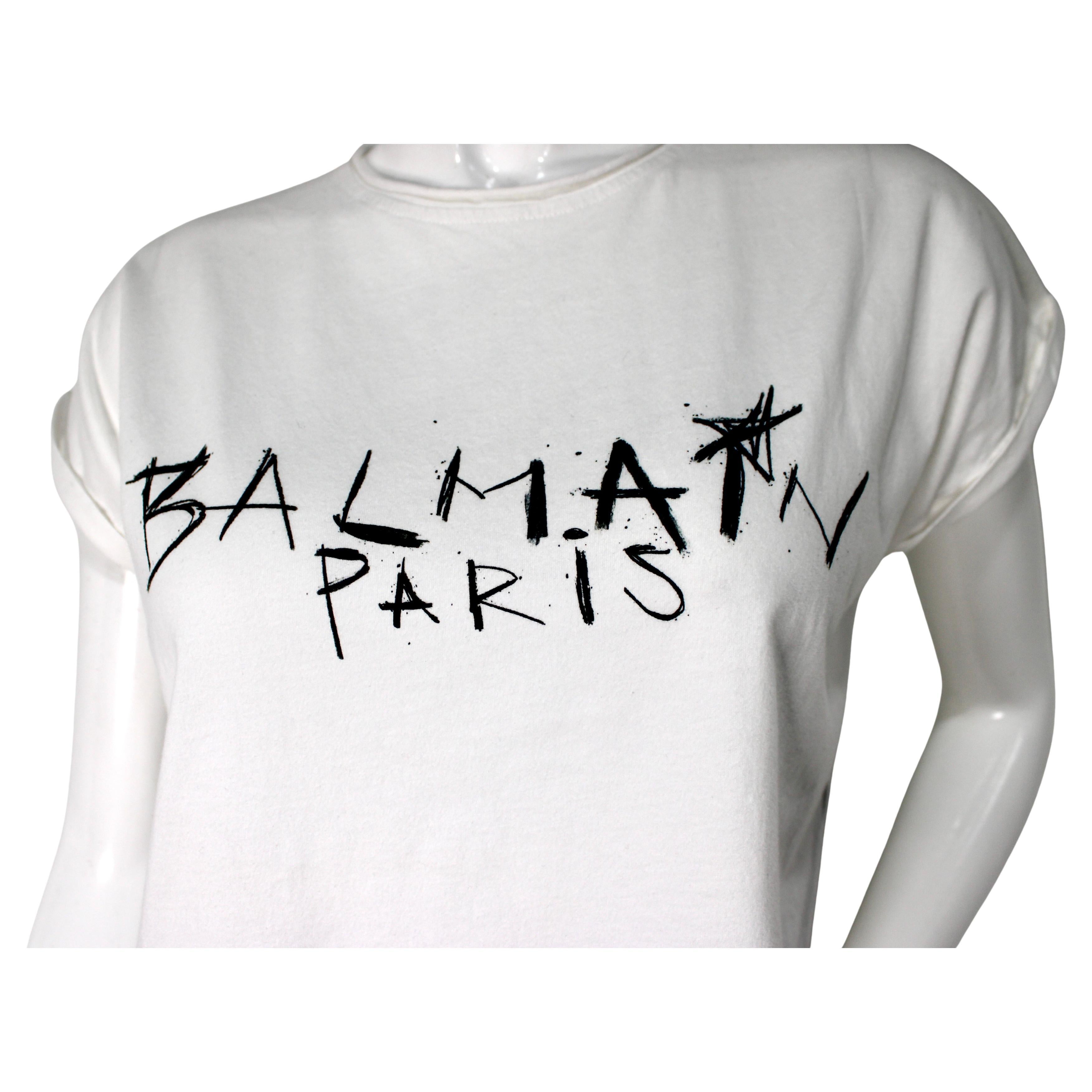 Balmain White & Black Graffiti Style Cotton Cropped T-Shirt ist ein gewagtes und modernes Stück, das urbane Streetwear mit hochwertiger Designerkunst verbindet. Das aus Baumwolle gefertigte T-Shirt ist mit einem auffälligen, von Graffiti