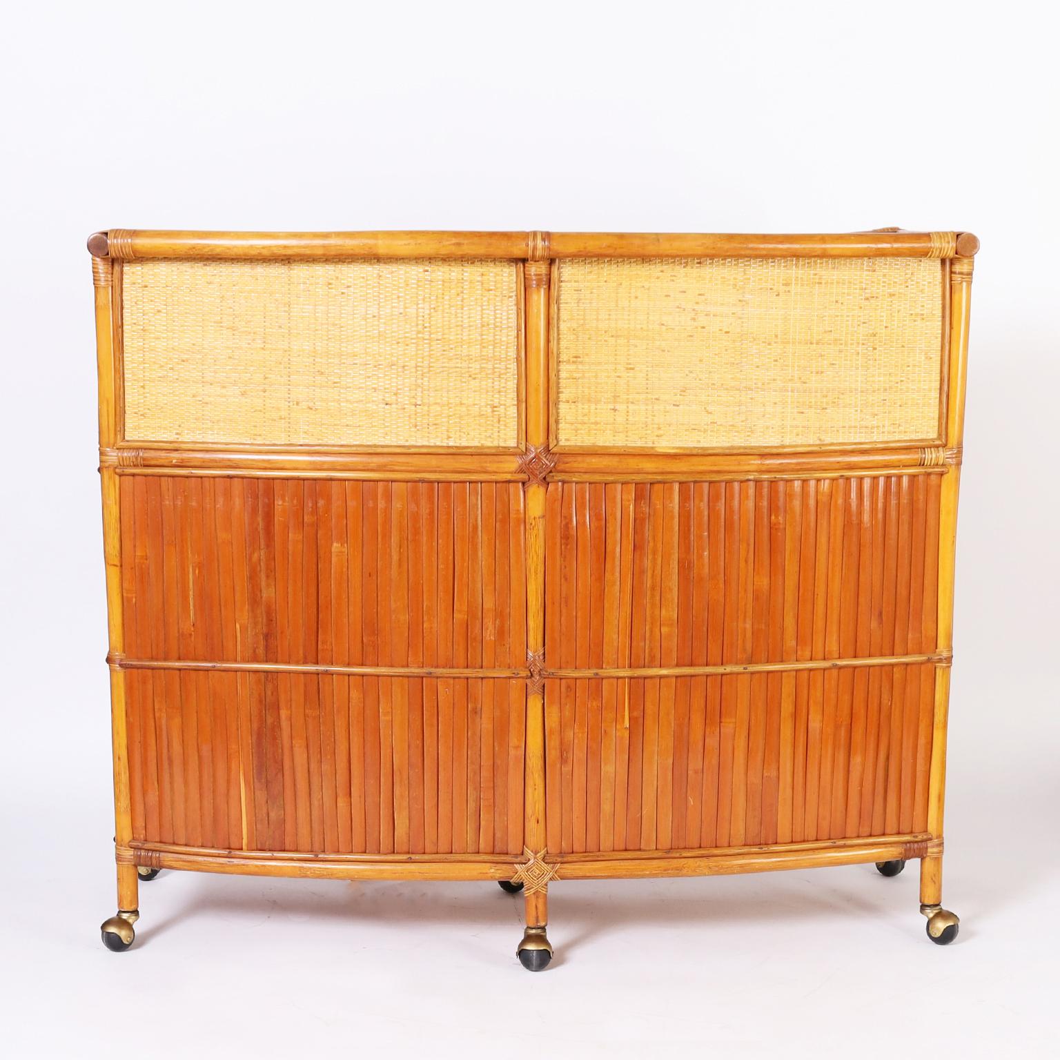 In vogue vintage demilune serving bar crafted with a bamboo frame having grasscloth panels on the top and sides, reed wrapped joints, service area with plenty of storage and set on rolling casters. 