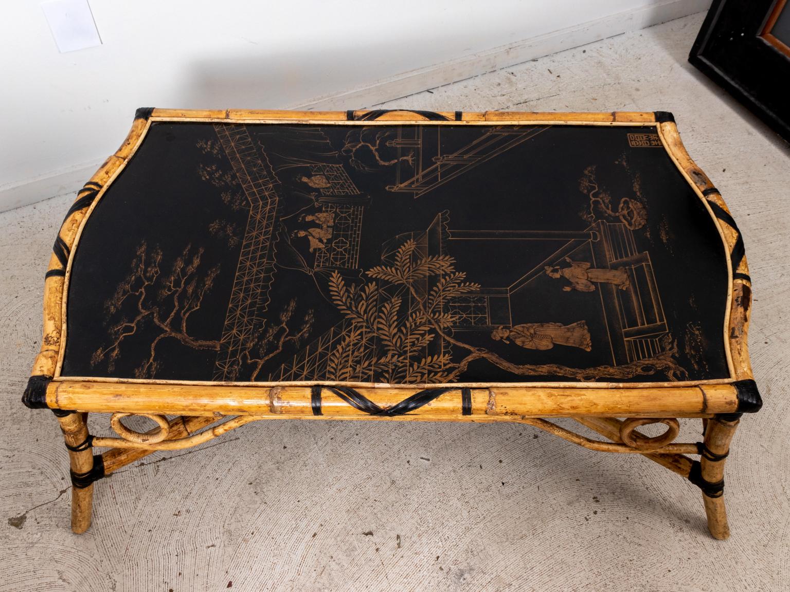 Bamboo frame coffee table with black and gold painted Chinoiserie decoration on the tabletop. The panel on the tabletop depicts Chinoiserie style figures in a village scene with trees. The table itself is accented with circular medallions on the