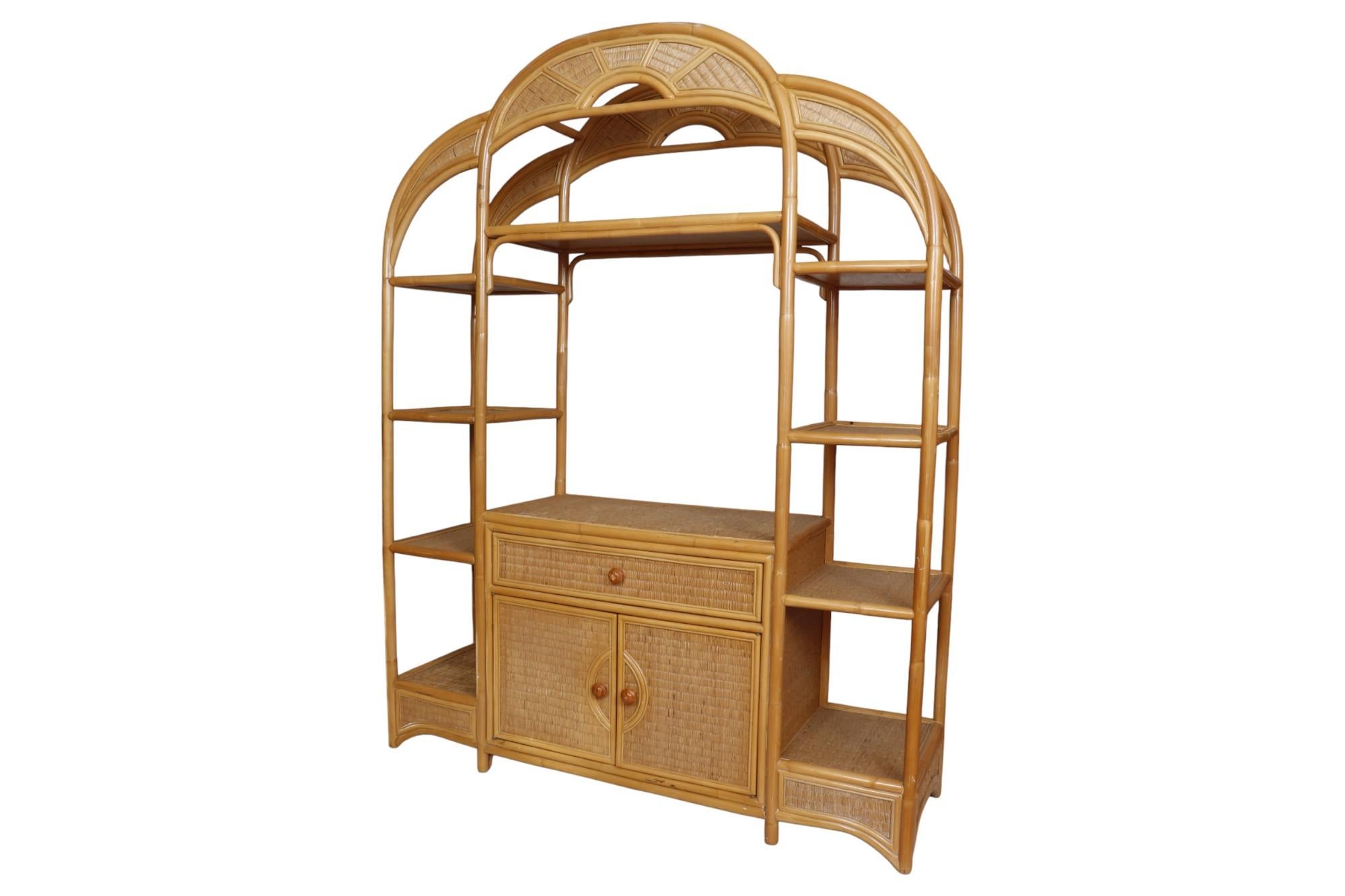 A bamboo and rattan etagere. The bamboo frame forms three arches, the central arch is pierced and decorated with a sunburst motif. Nine open shelves flank a central draw and cabinet. Both open with wooden mushroom shaped knob handles.