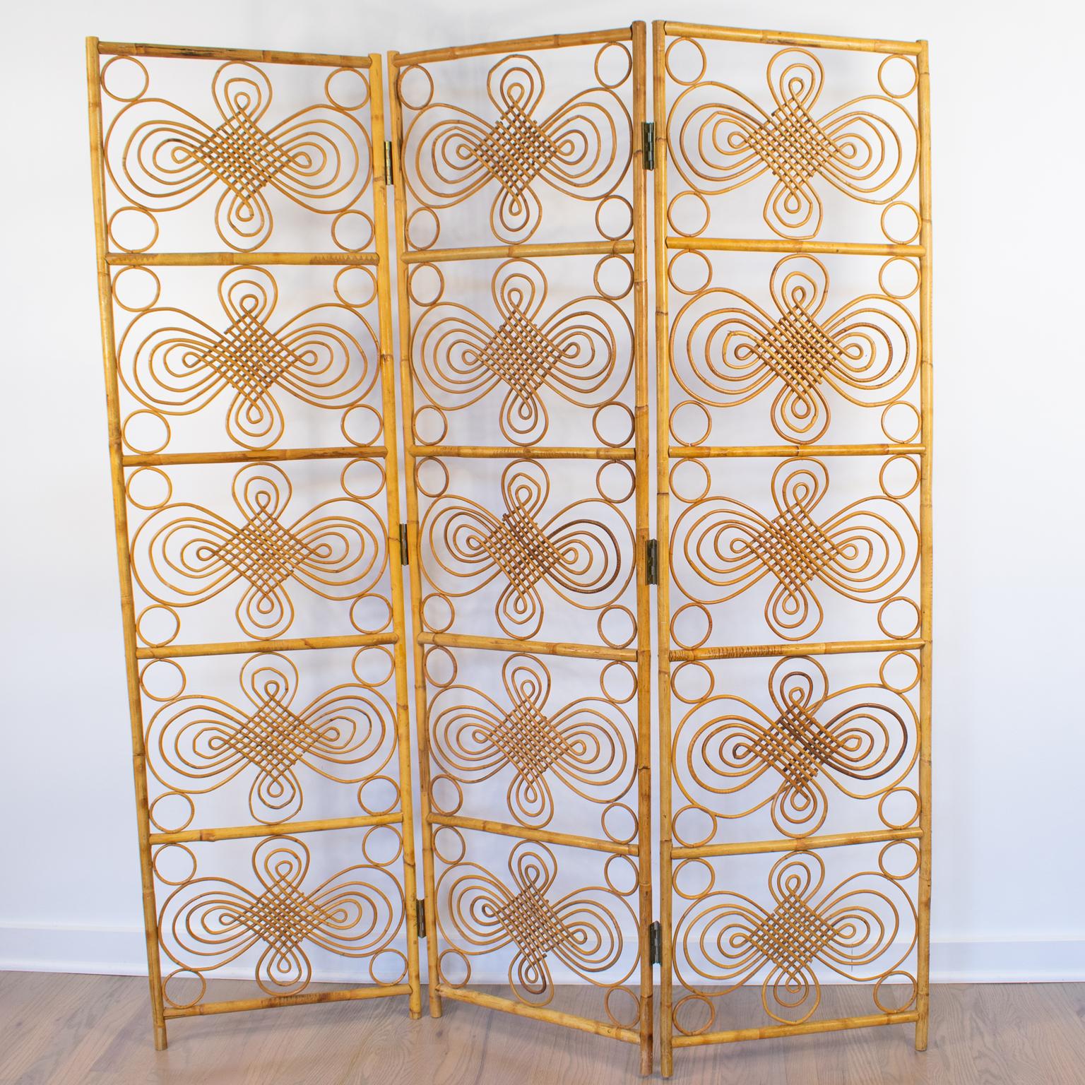Hand-crafted in France in the 1960s, this striking rattan or wicker and bamboo three-panel room divider boasts a typical Mid-century filigree decoration. This rattan screen features mid-century and chinoiserie accents, circular ornaments, and a