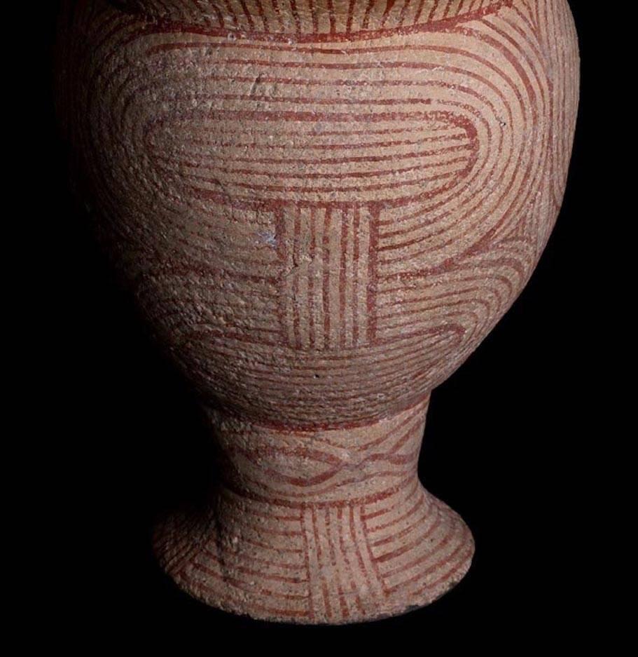 A pottery vase having an egg shape body with a flared rim, standing on a pedestal base. Cream ground with an intricately painted red geometric linear decoration. Mineral deposits on the surface. 

Condition: Professionally restored from