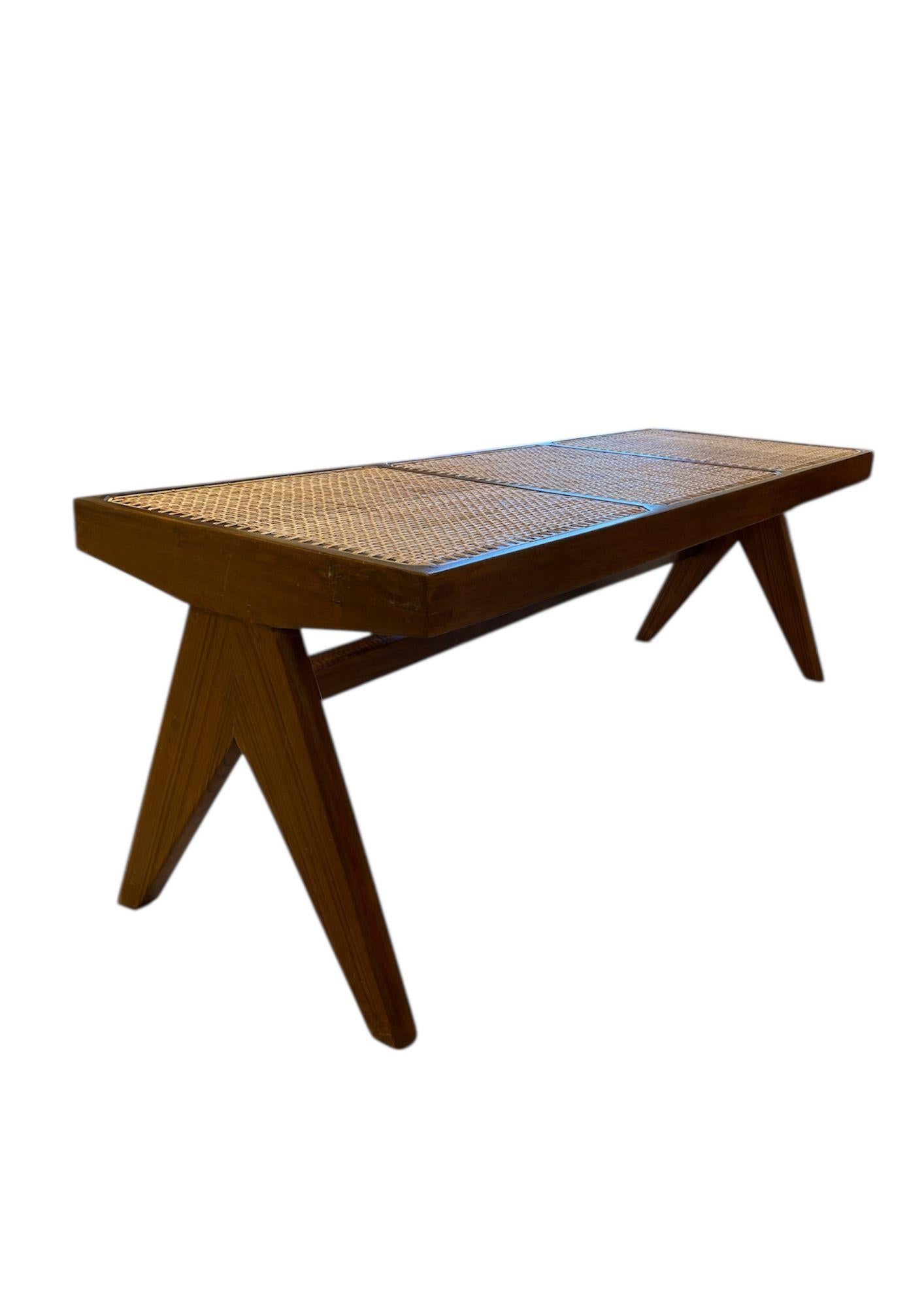 Solid teak bench with woven cane seating by French designer Pierre Jeanneret. Rectangular seat resting on two “compass”-style side legs connected by a low H-shaped crossbar.