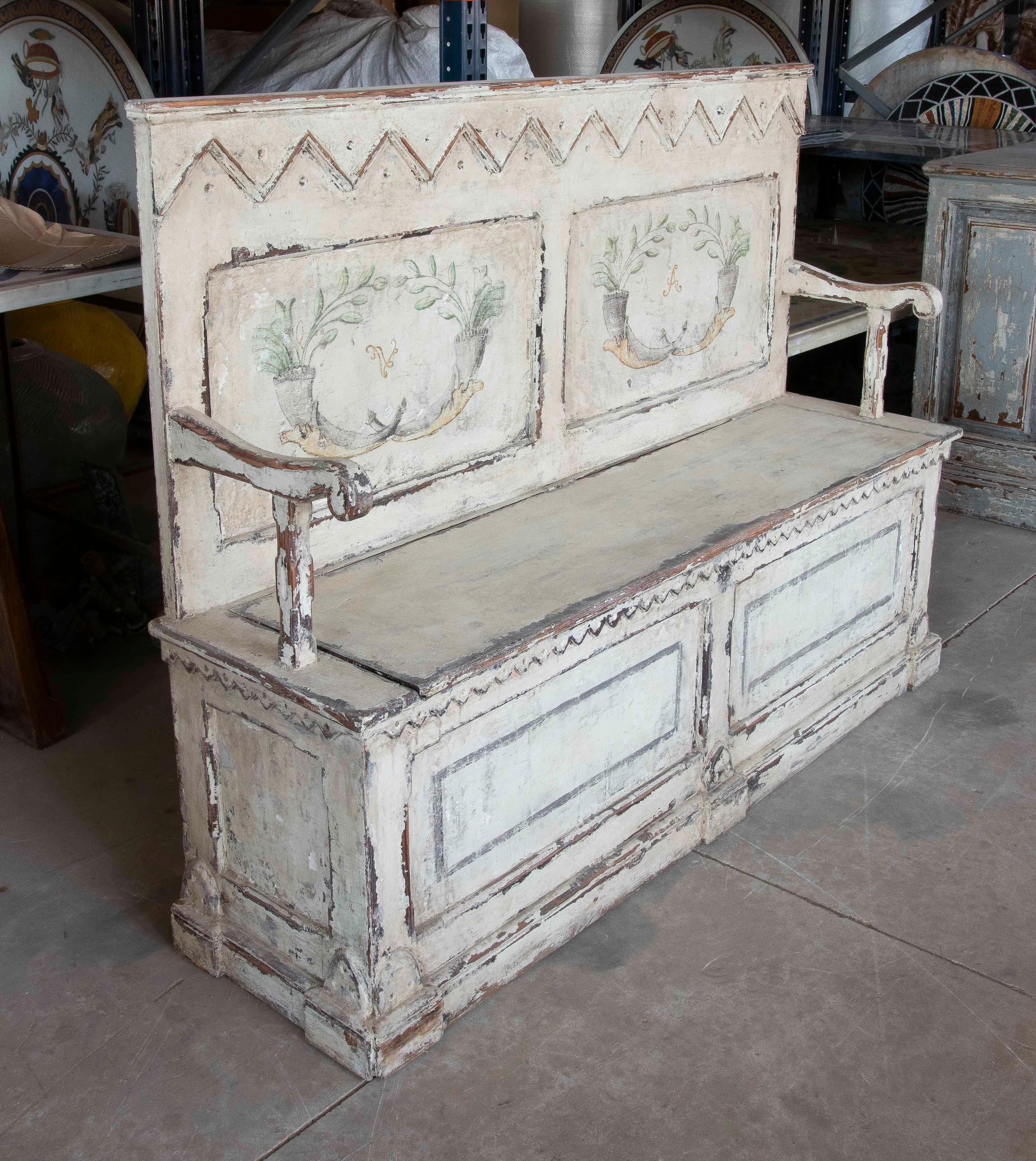 Charming wooden bench painted in aged white tones, decorated with hand-painted floral motifs on the backrest. The fold-down seat reveals a practical trunk-style storage space, ideal for entryways, living rooms, or relaxation areas with a Provençal