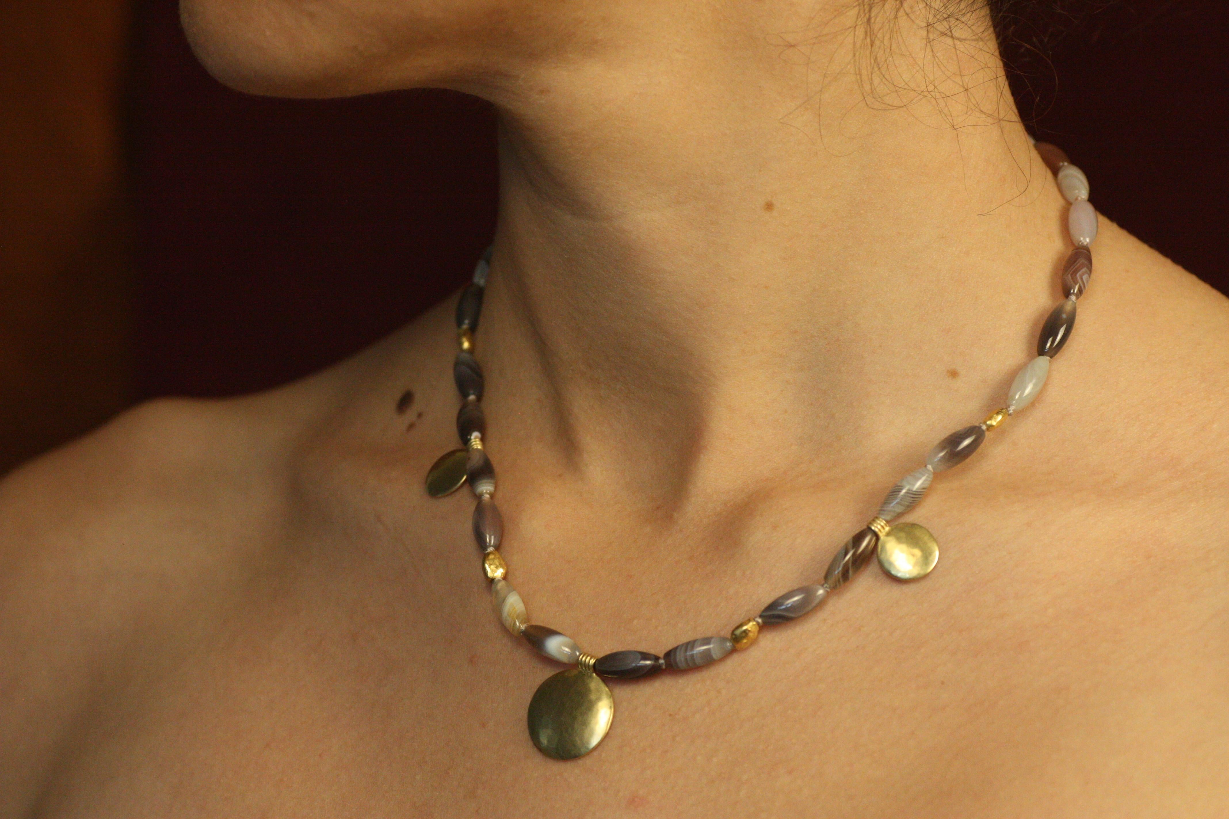 Questa collana di dischi solari, unica nel suo genere, abbina la bellezza terrosa dell'agata a bande con le pepite d'oro ad alta caratura e i dischi solari. Su ogni perlina di colore grigio danzano vortici e bande uniche e naturali, alcune chiare e