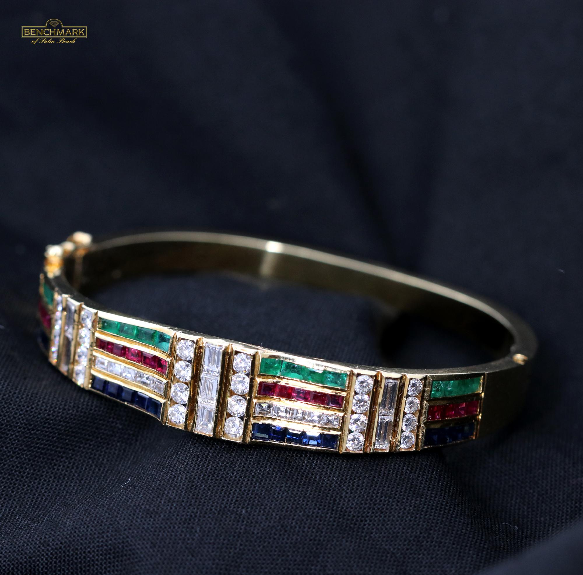 An 18K yellow gold hinged cuff bracelet measuring 1/2 an inch at the center, and 3/16 of an inch at the base. At the center of the bracelet are three baguette cut diamonds, from which extend alternating rows and columns of diamonds, emeralds,