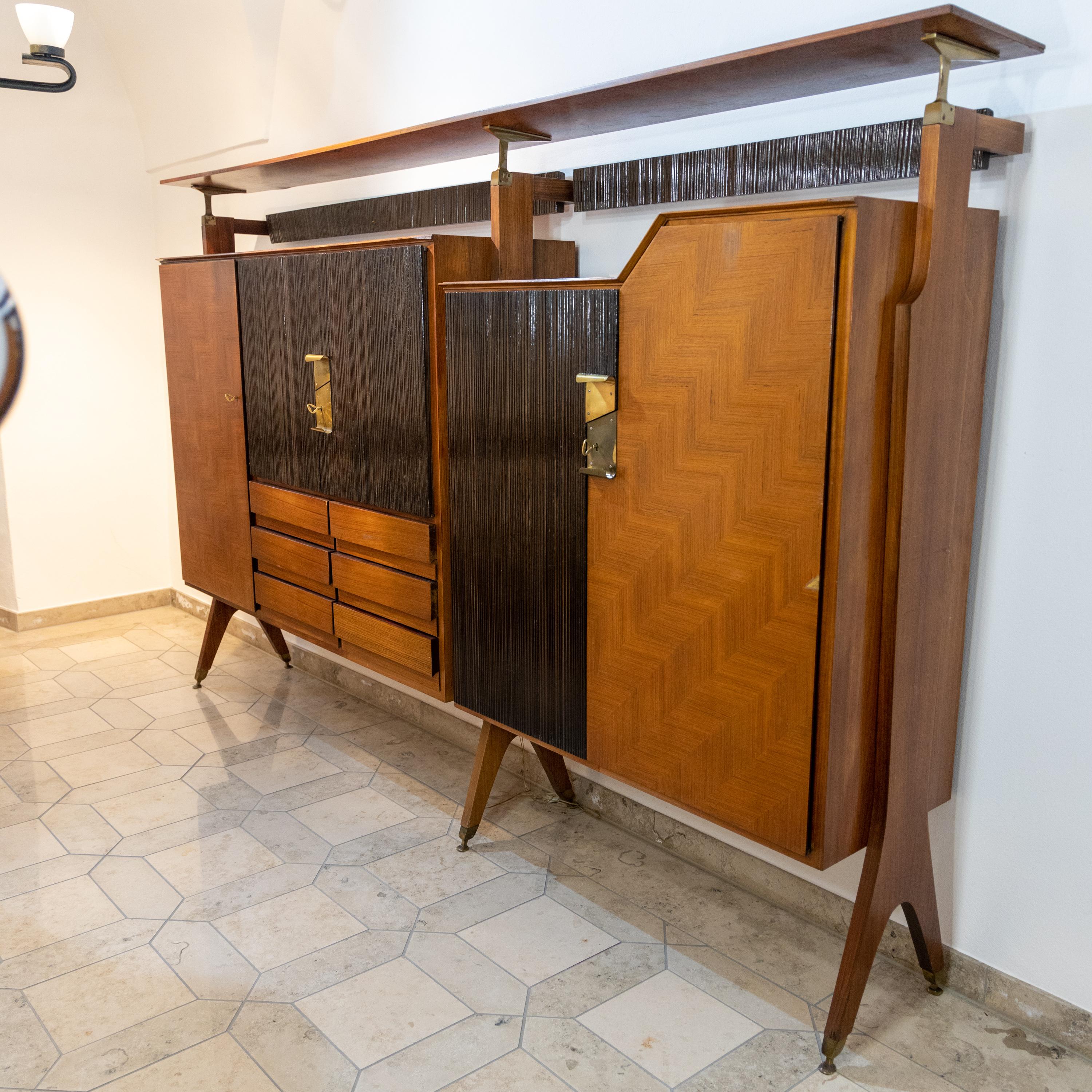 Bar cabinet standing on forked legs with asymmetrical doors and grooved or veneered surfaces and a continuous shelf on top.