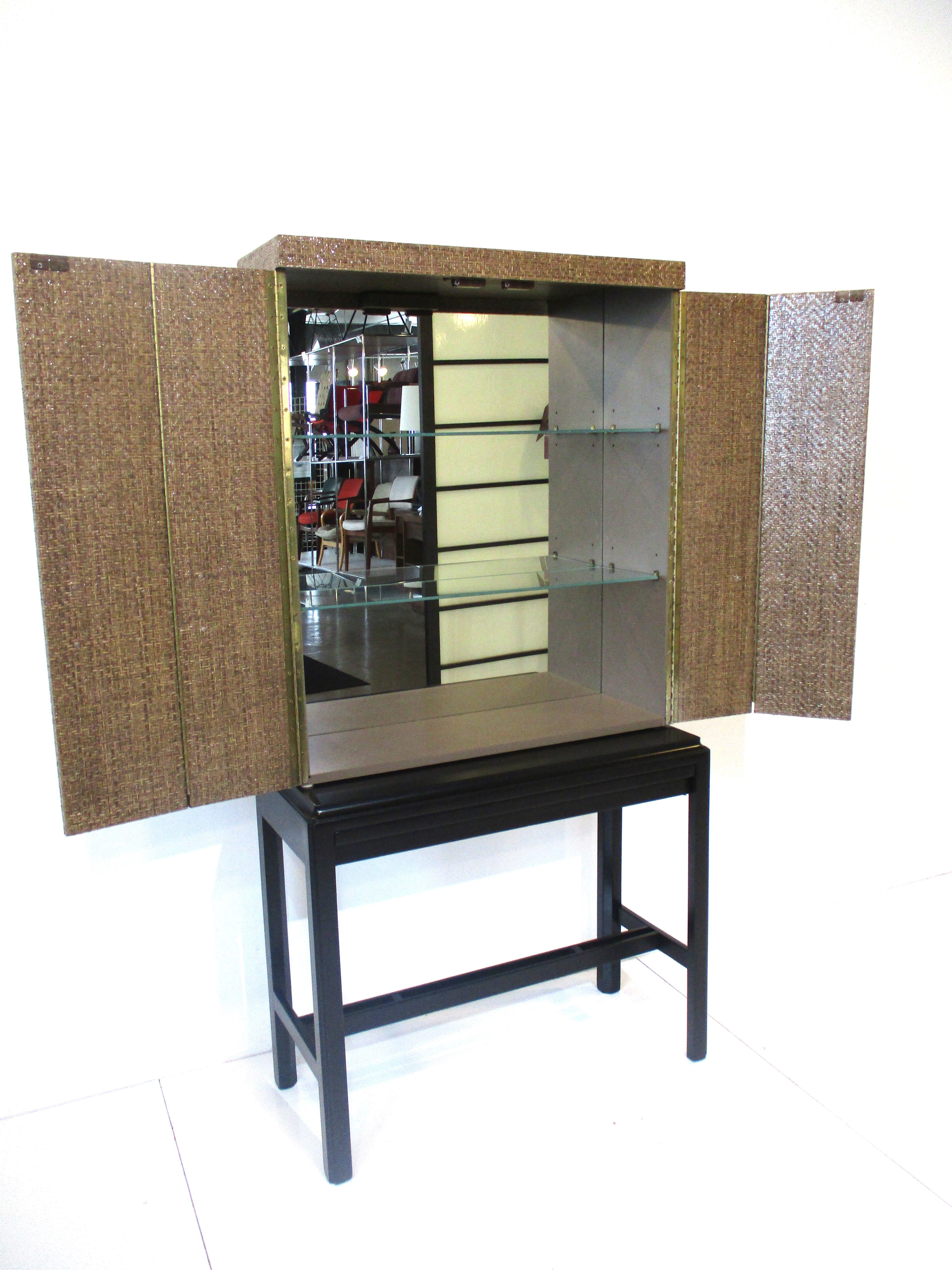 A very well crafted two piece bar cabinet with sea grass cloth lacquered top cabinet and bi folding doors having brass piano hinges with brushed brass / bronze pulls . Inside are two adjustable glass shelves and mirror backing making for a bright